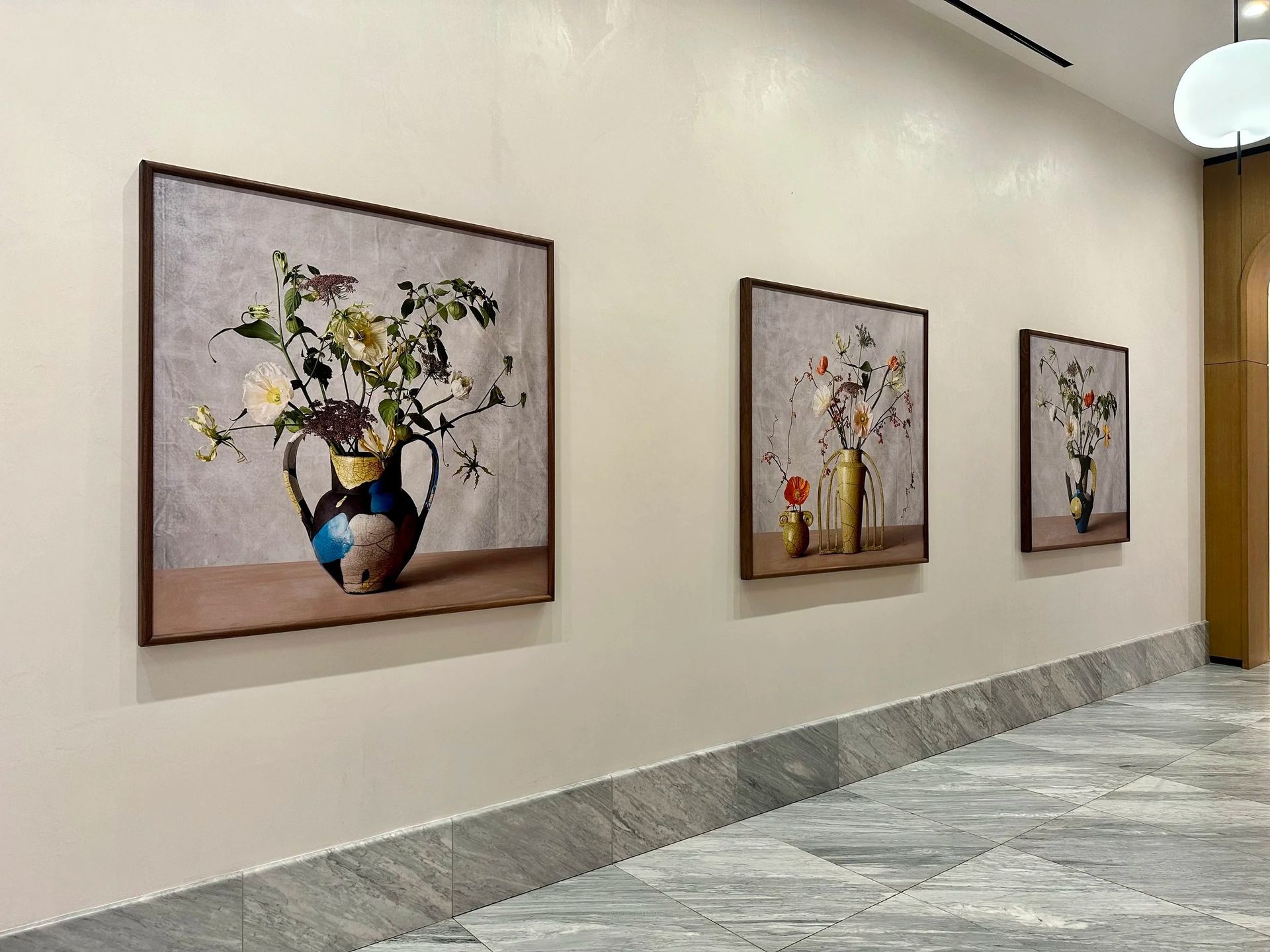 Three framed flower still-life paintings hang on a beige hallway wall above a gray tiled floor.