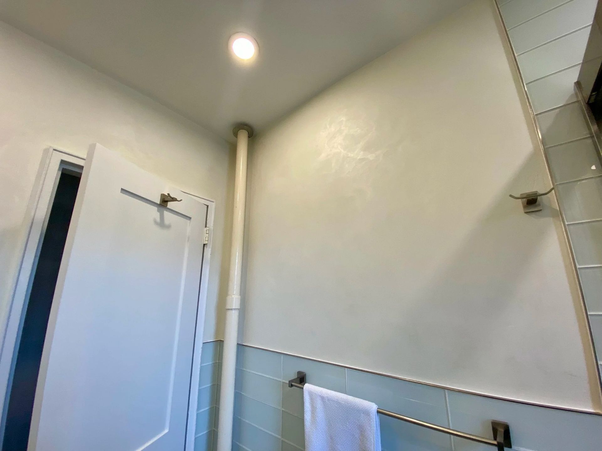 Bathroom interior: light blue walls, white door, towel bar with towel, pipe, and overhead light.