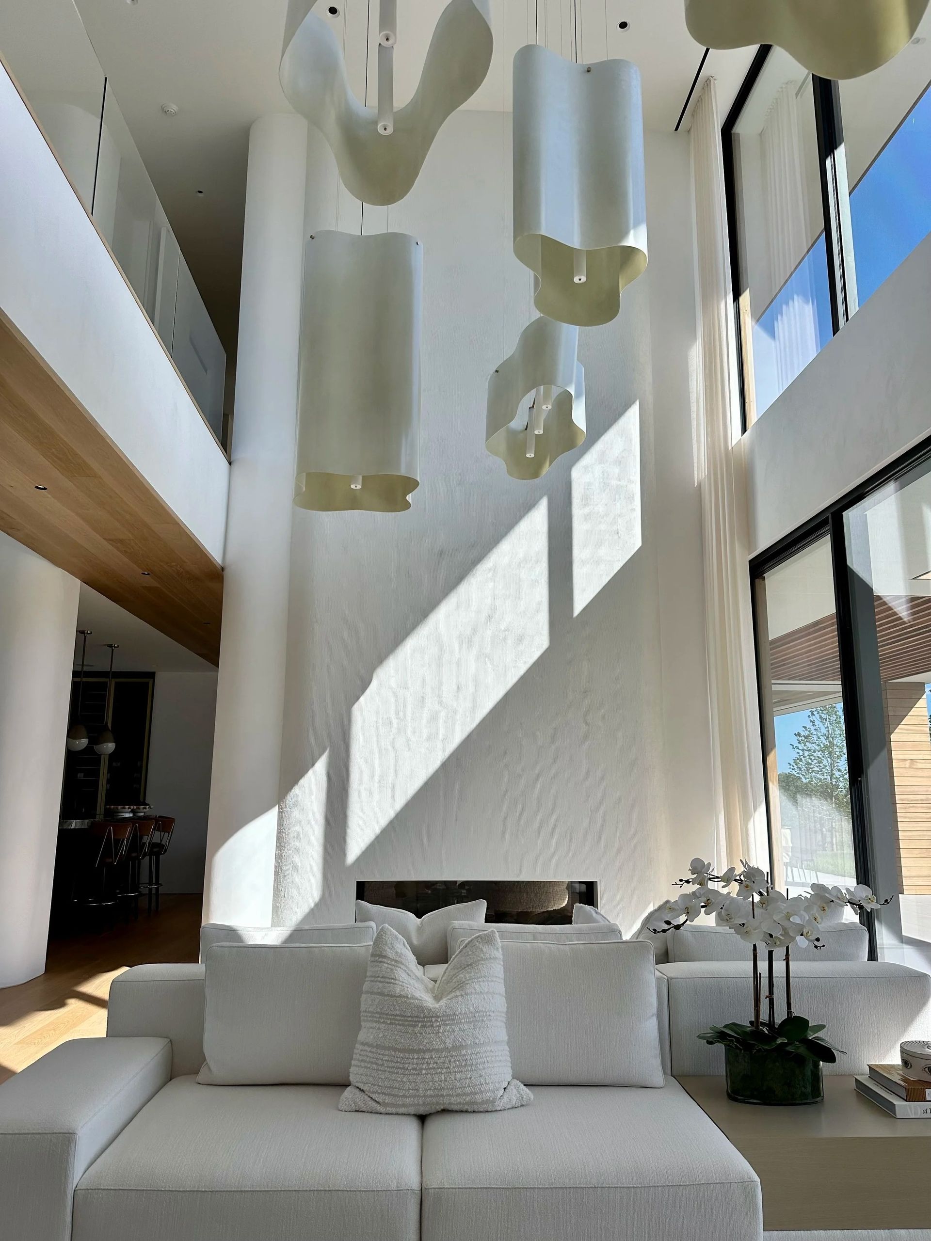 Modern white living room with high ceilings, large windows, and sculptural light fixtures above a white sofa.