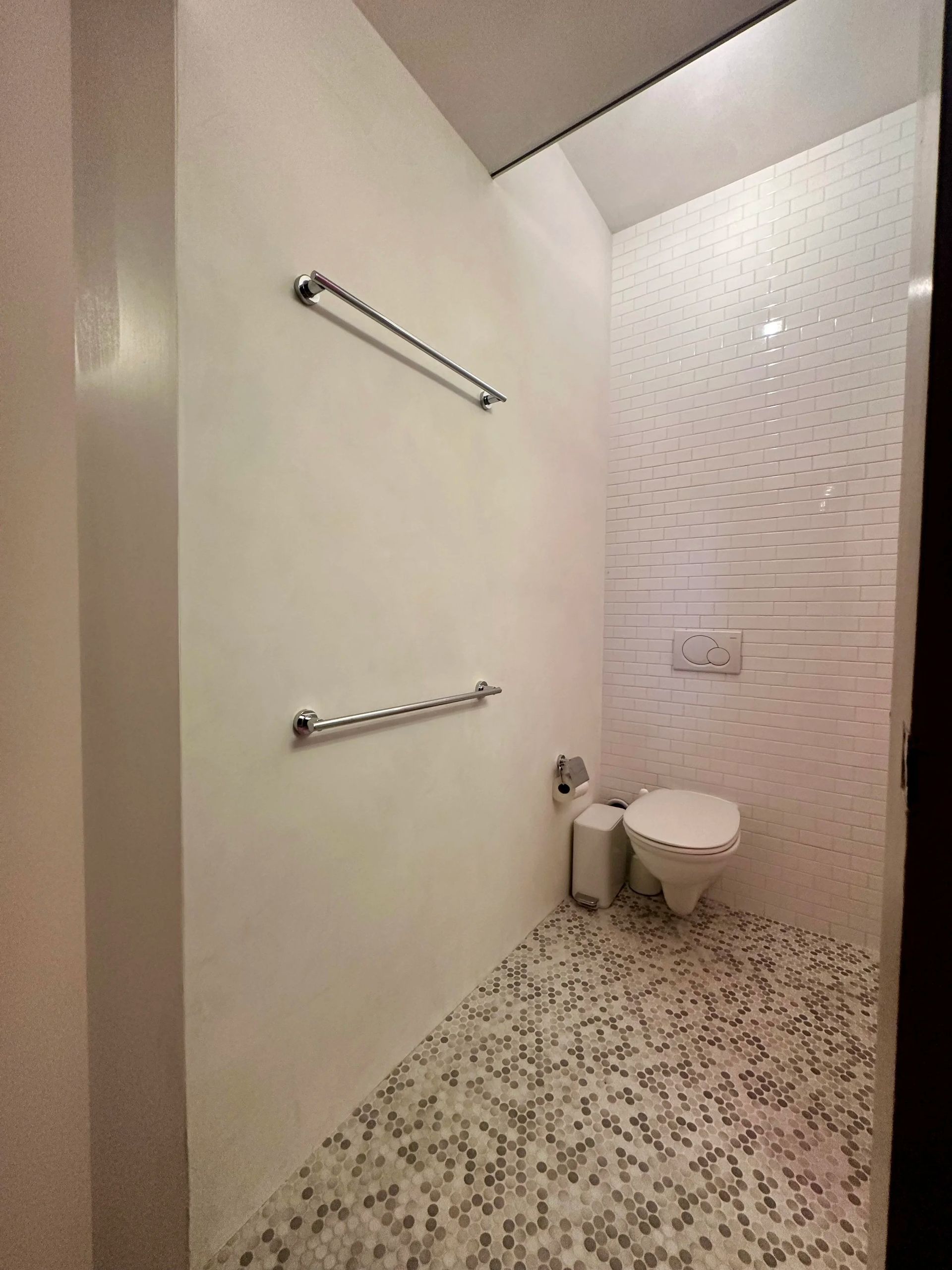 Small, tiled bathroom with toilet, white walls, and silver towel racks.