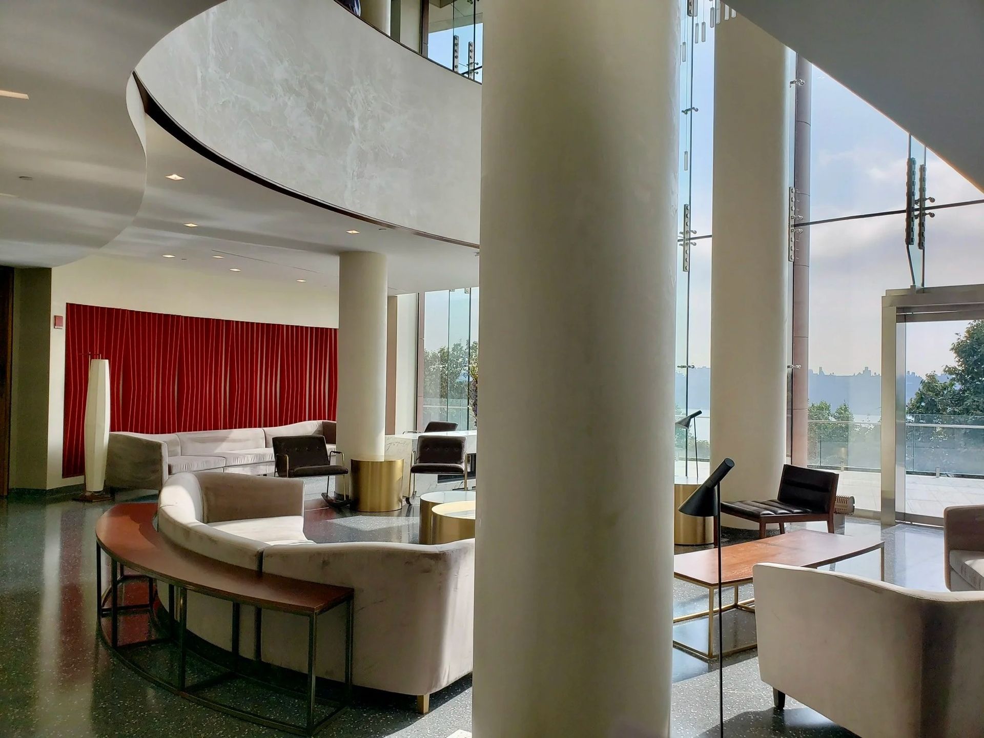 Lobby with large white columns, curved seating, red curtains, and floor-to-ceiling windows overlooking a body of water.