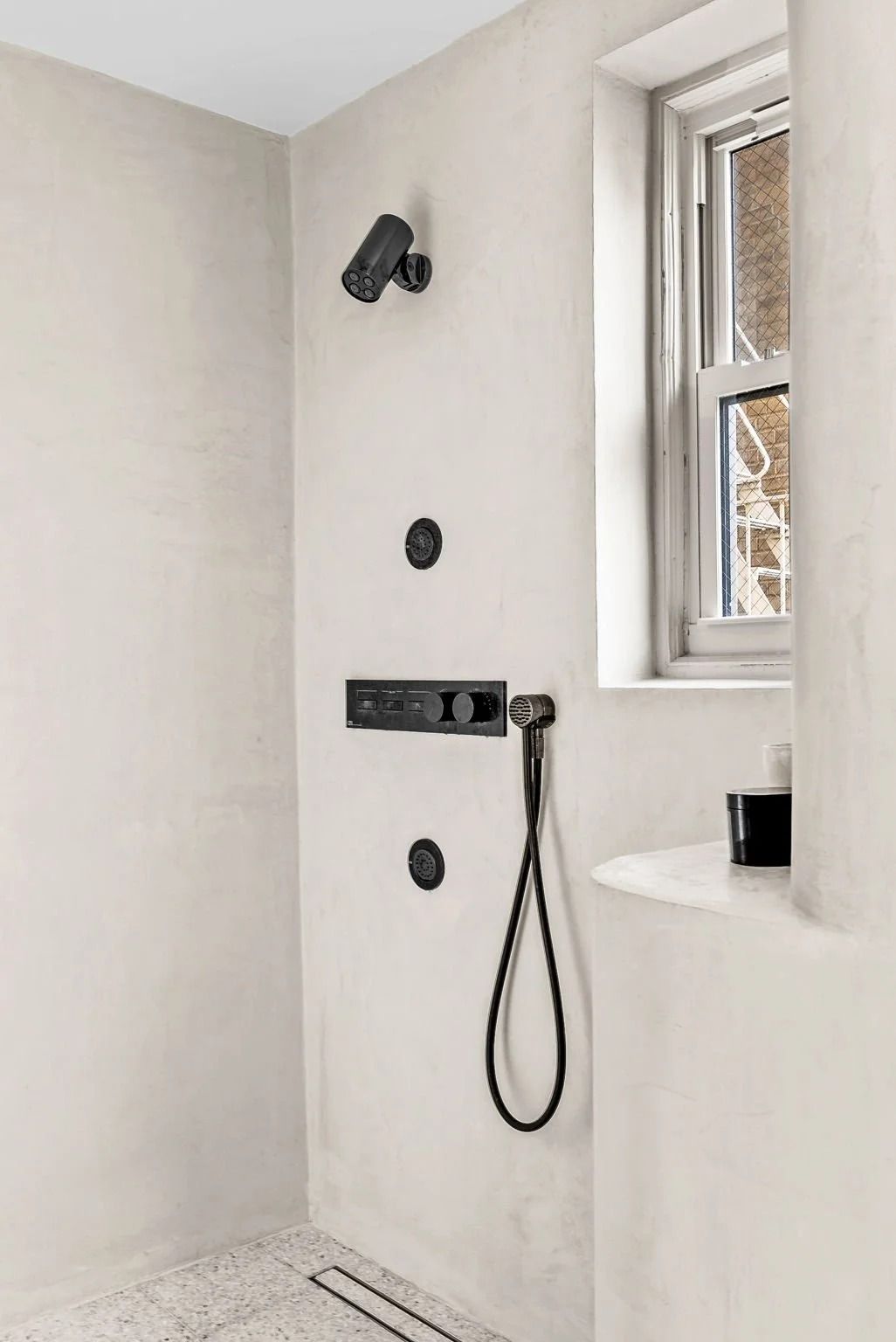 Shower with black fixtures against a light-colored, textured wall near a window.