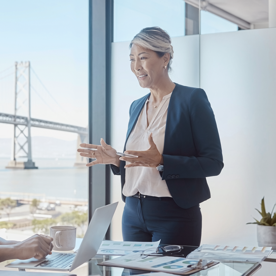 Businesswoman presenting in a bright office with a bridge view, holding a tablet beside a laptop and coffee mug