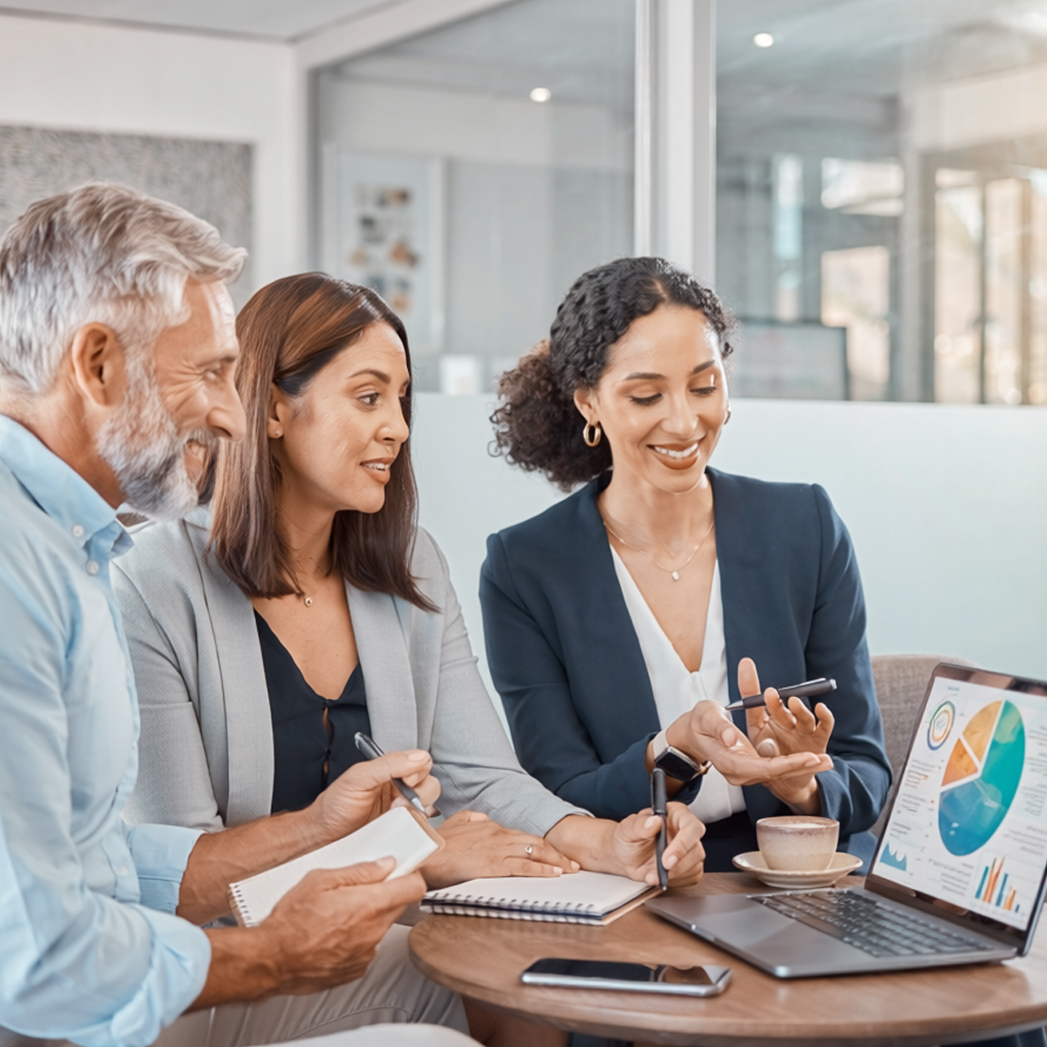 Three coworkers discuss a laptop chart during a meeting in a glass-walled office.
