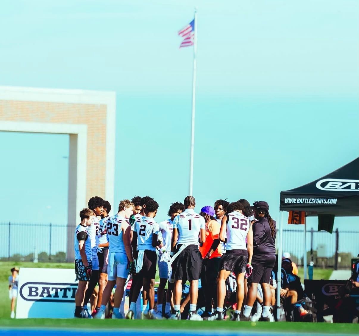 Youth sports team huddled on a field near a tent and flagpole under a clear blue sky