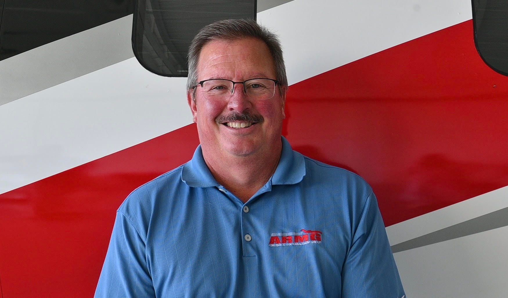A man wearing glasses and a blue shirt is smiling in front of a red and white striped wall.
