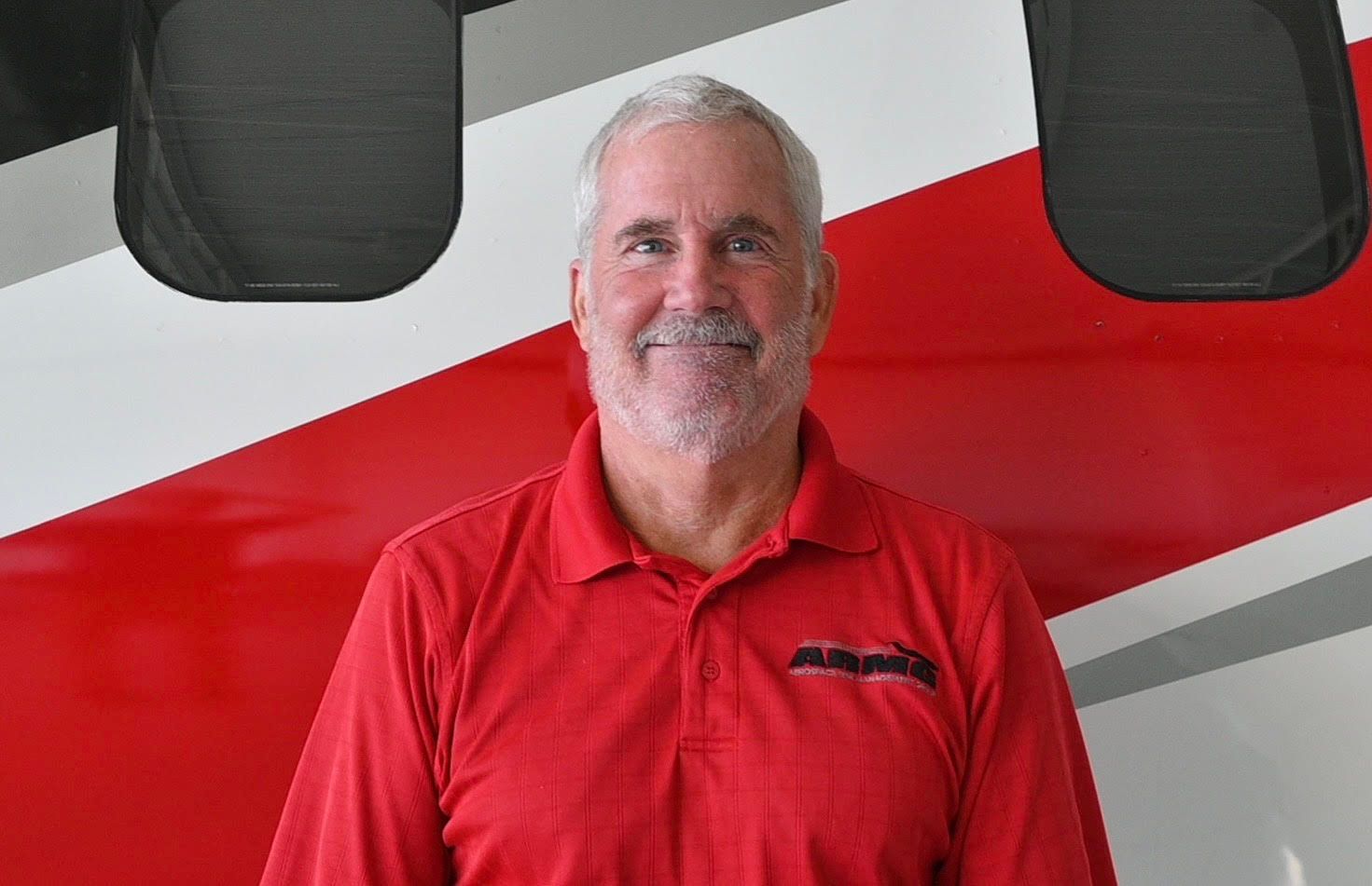 A man in a red shirt is smiling in front of a red and white airplane.