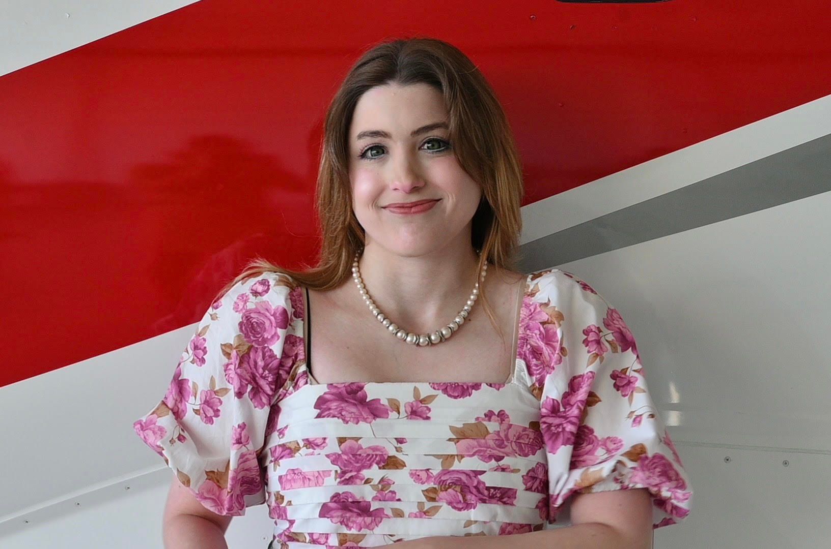 A woman in a floral dress and pearl necklace is standing in front of a red and white wall.