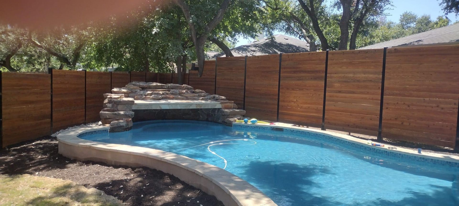 A large swimming pool with a wooden fence and a waterfall in the backyard.