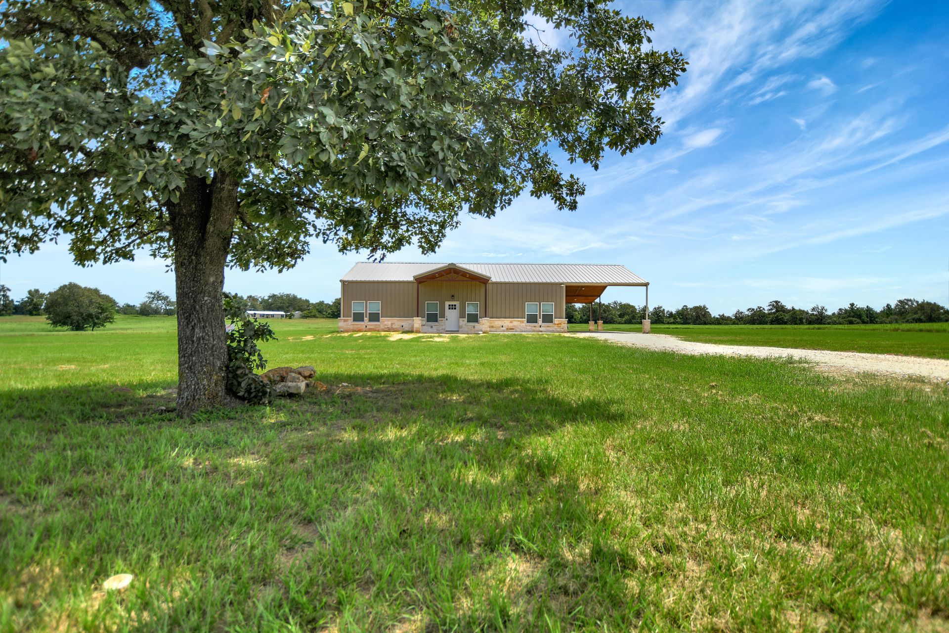 A single-story tan house with a porch sits in a grassy field, a large tree in the foreground.