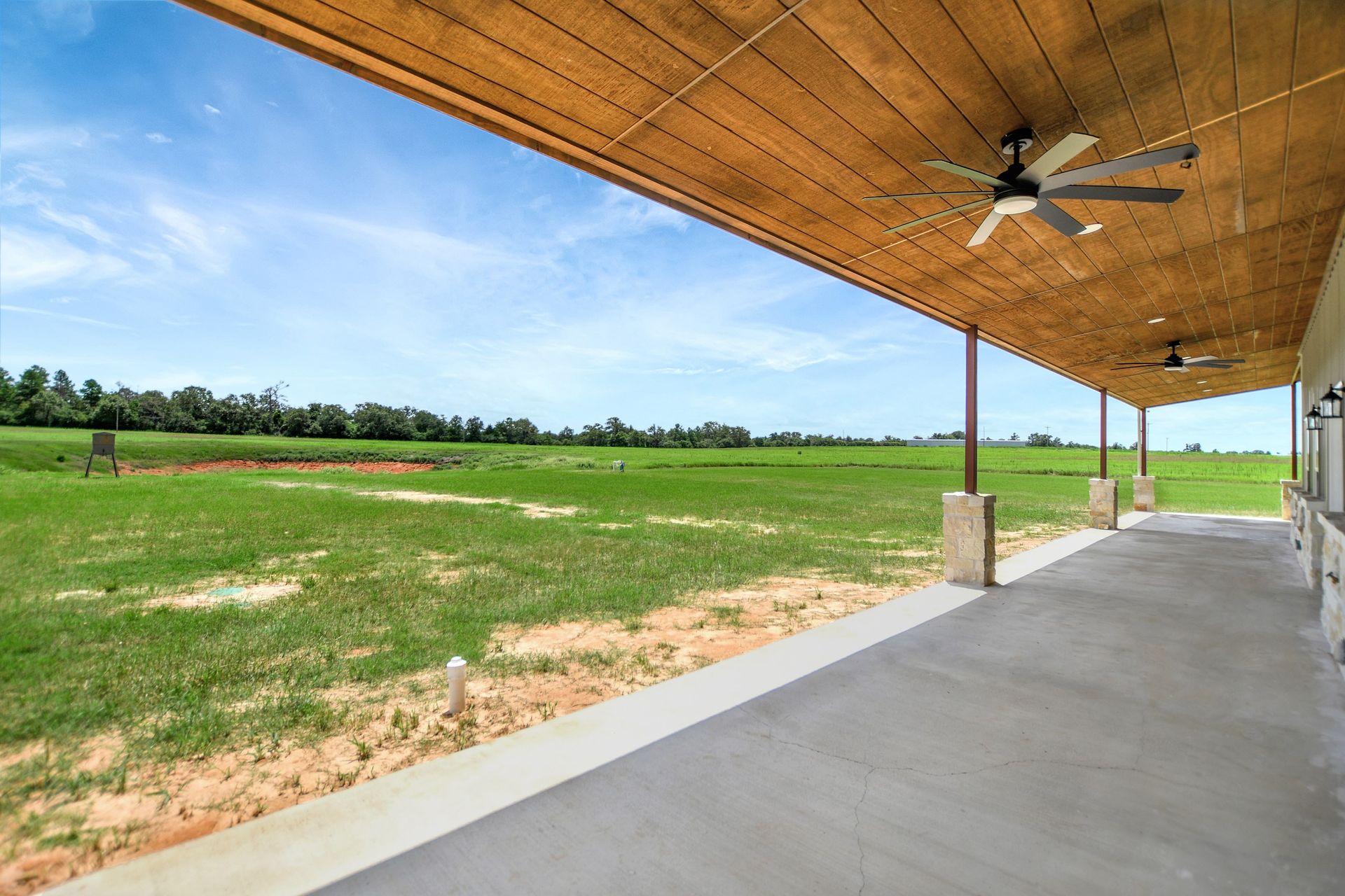 Covered patio overlooking a grassy field under a blue sky. Wooden ceiling, concrete floor, and ceiling fans.