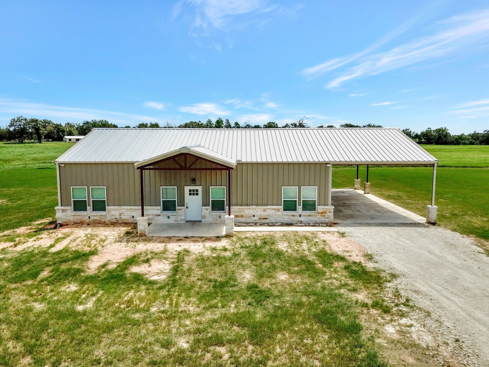 Tan metal-sided house with carport, white door, and green field under a blue sky.