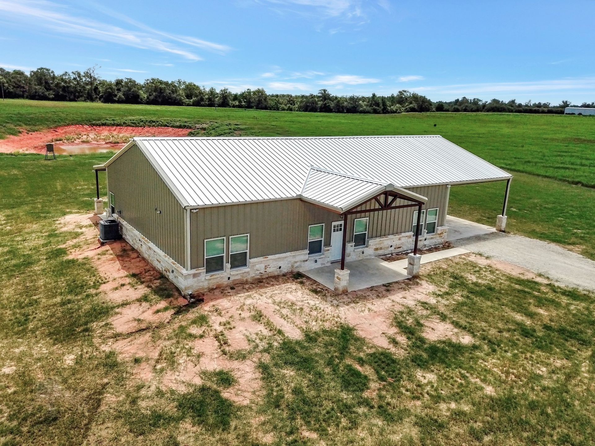 Metal-roofed home with porch and gravel driveway on a grassy field under a blue sky.