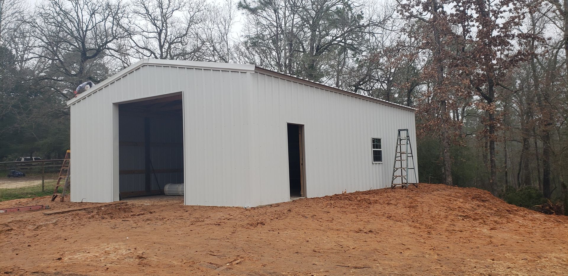A white metal building under construction on a dirt lot, surrounded by trees.