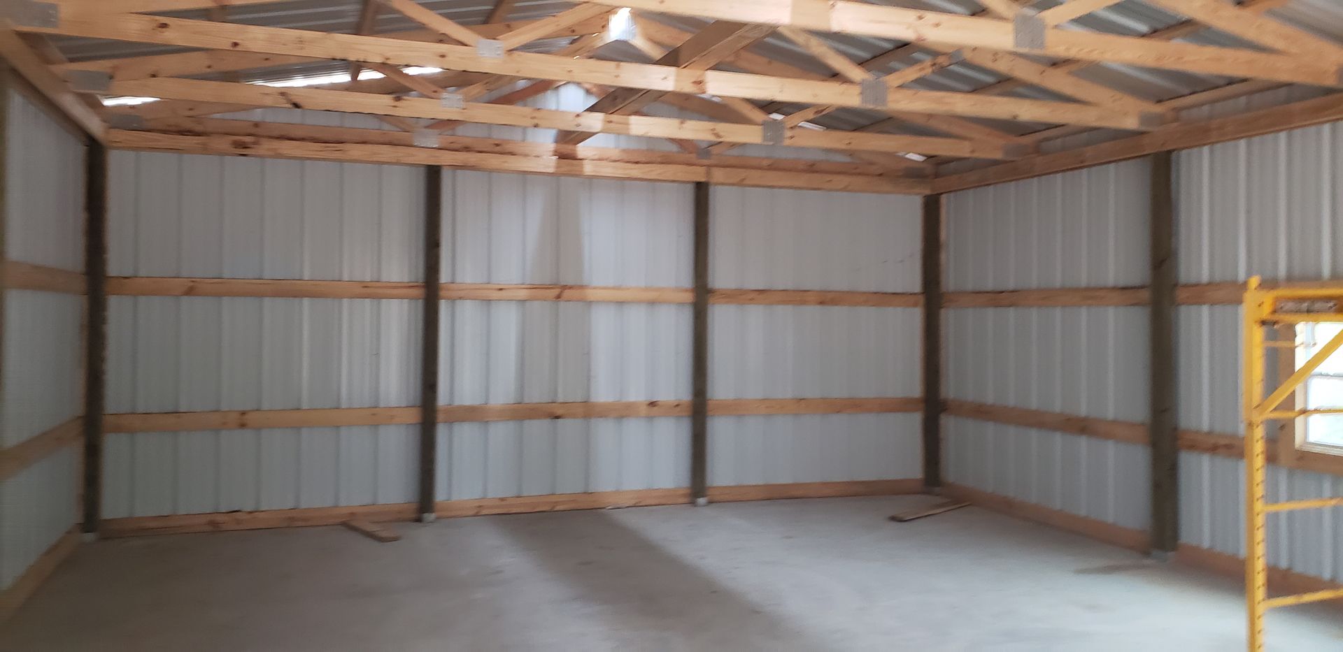 Interior view of a bare metal building under construction, with exposed wooden beams and a concrete floor.