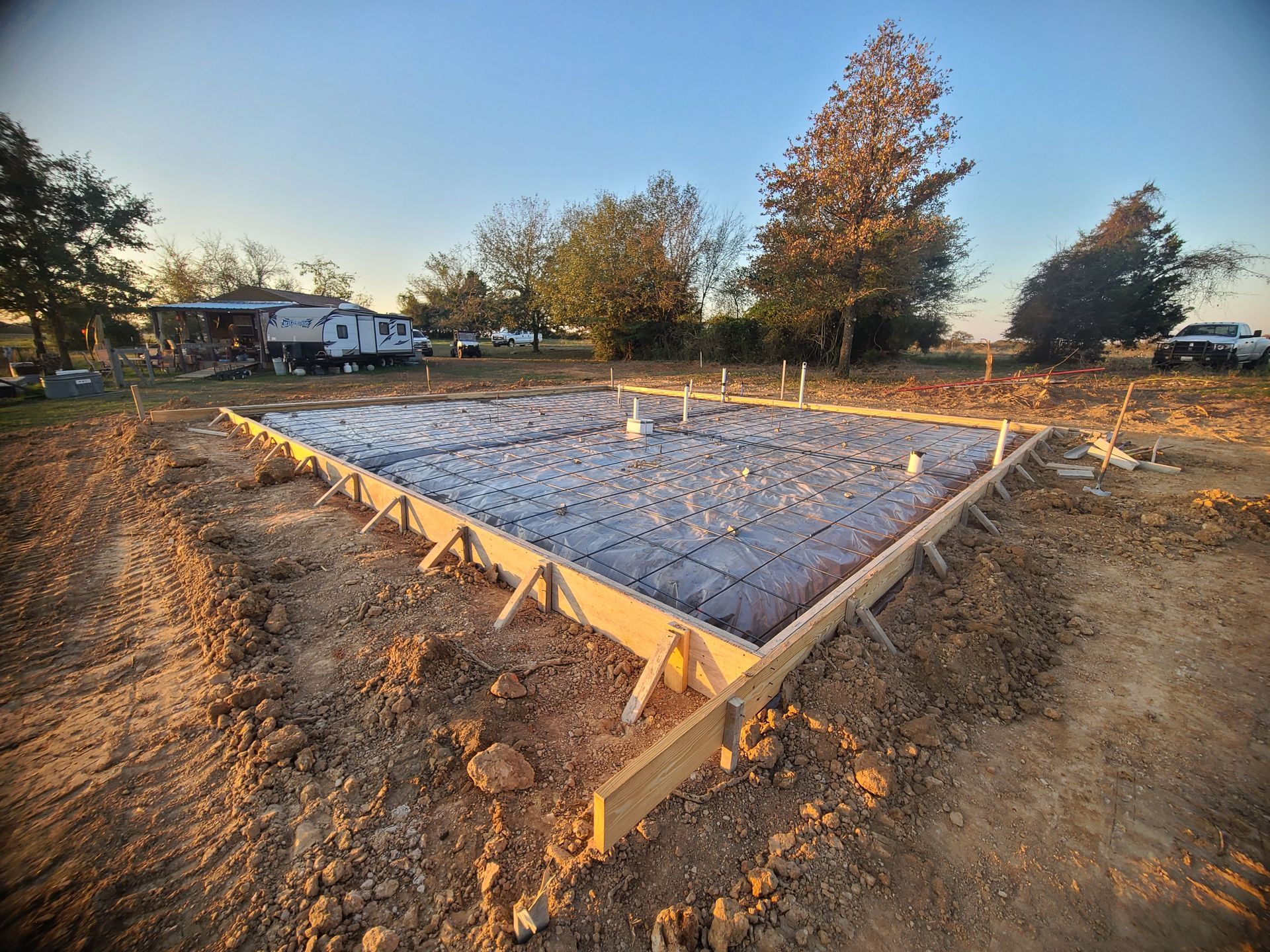 Newly poured concrete foundation within wooden frame, surrounded by dirt, with a trailer and trees in the background.