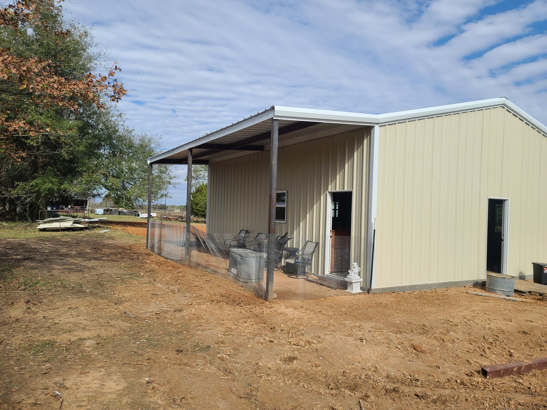 Beige metal building with a covered porch, a fenced area, and a dirt yard under a cloudy sky.