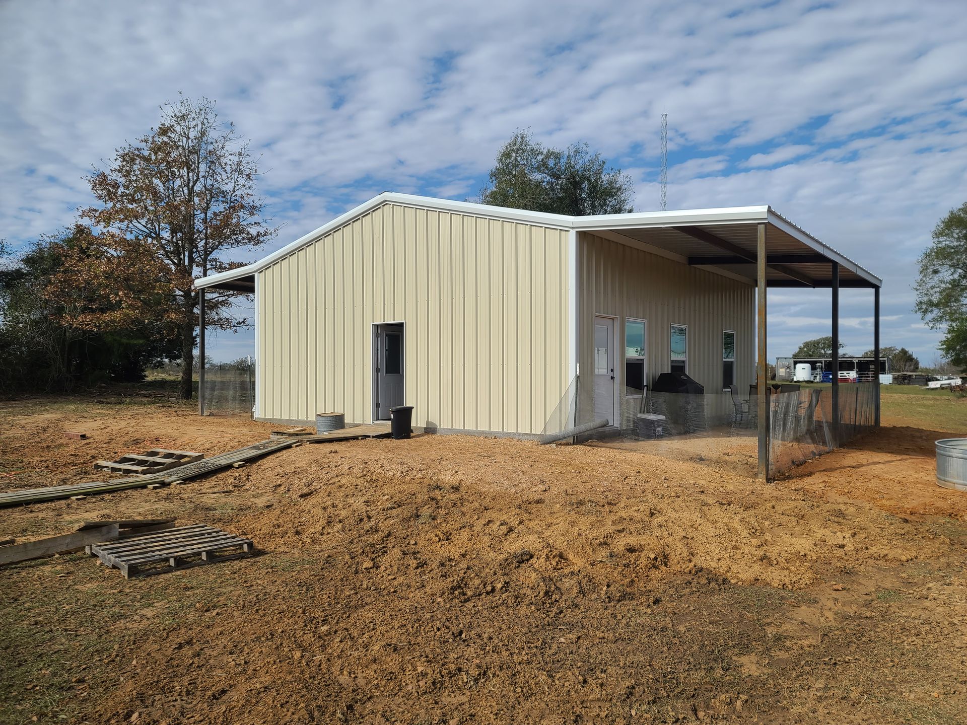 Beige metal building with a covered porch, set in a yard with a cloudy sky.
