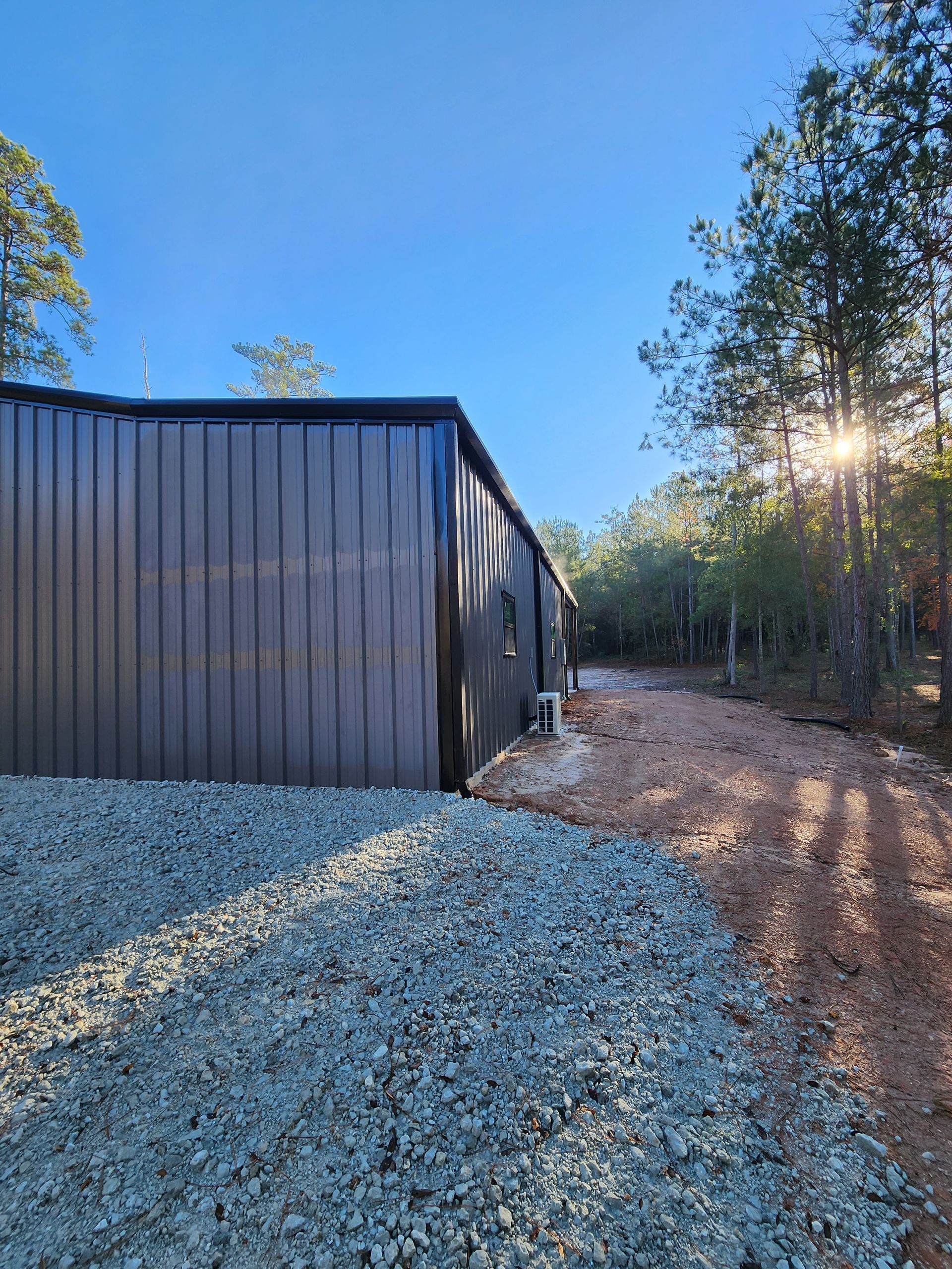 A dark metal shed with a gravel driveway set in a wooded area, with sunlight.