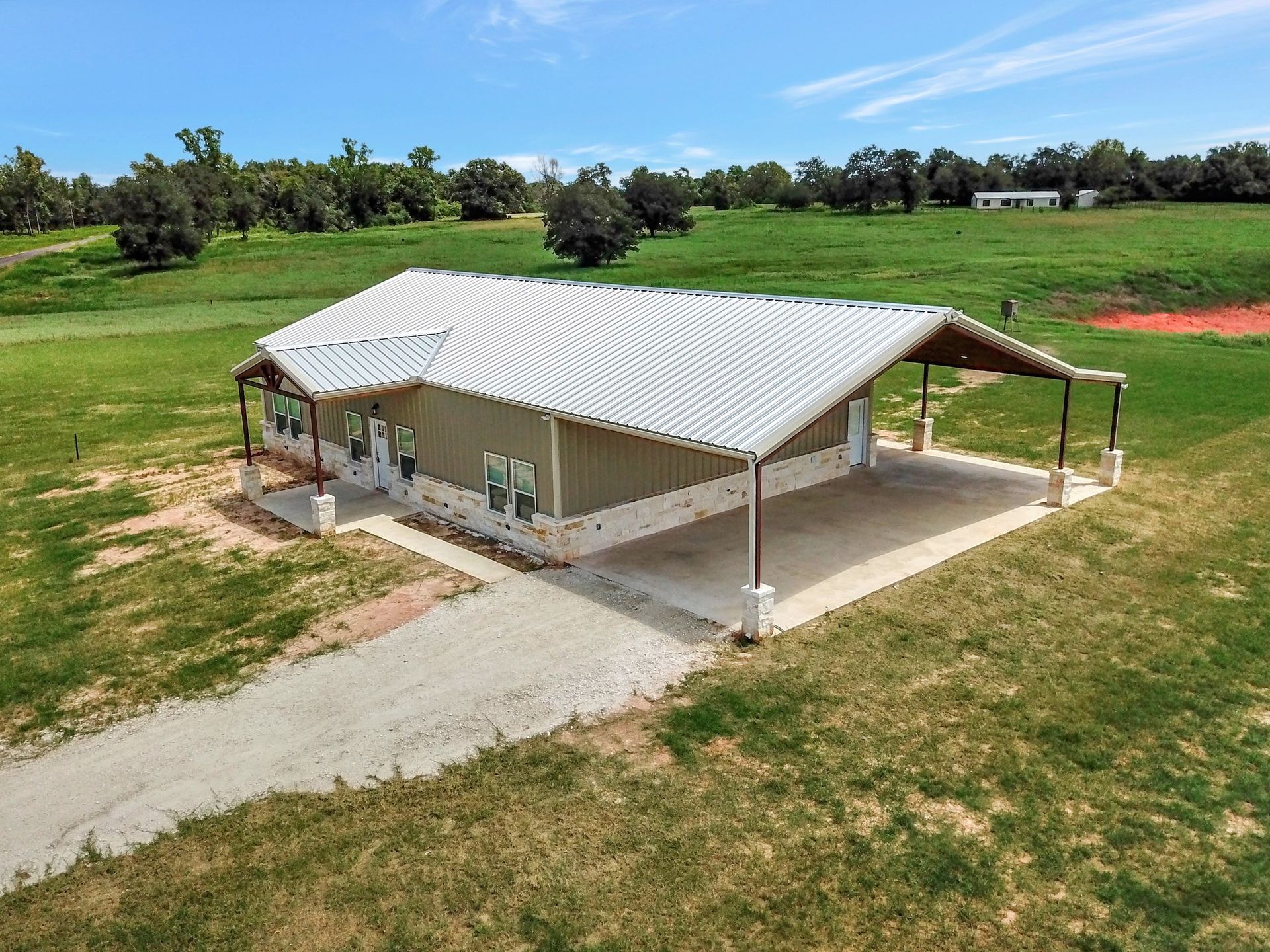 A light tan metal-sided house with a carport on green grassy property with a gravel driveway.