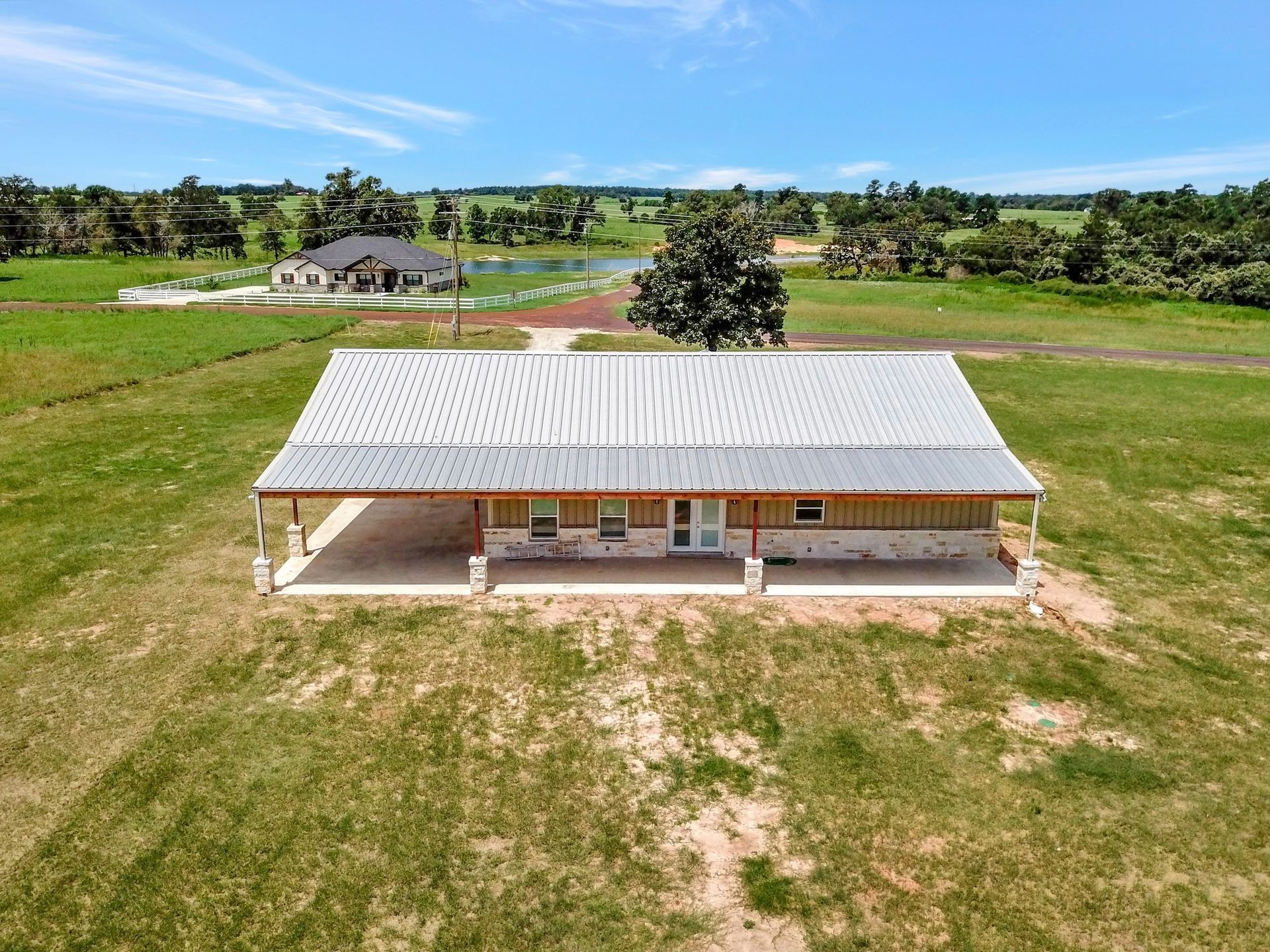 Drone view: single-story house with porch and metal roof on green field, another house and pond in distance.