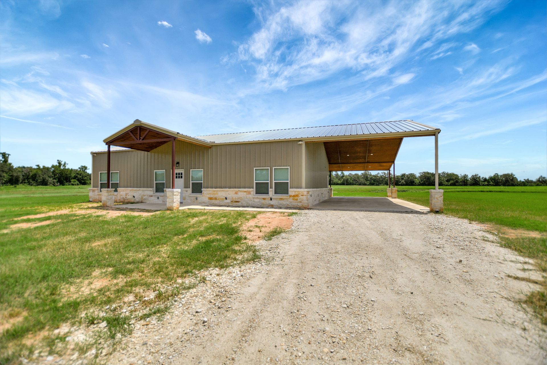Tan metal building with a carport, gravel driveway, and green fields under a blue sky.