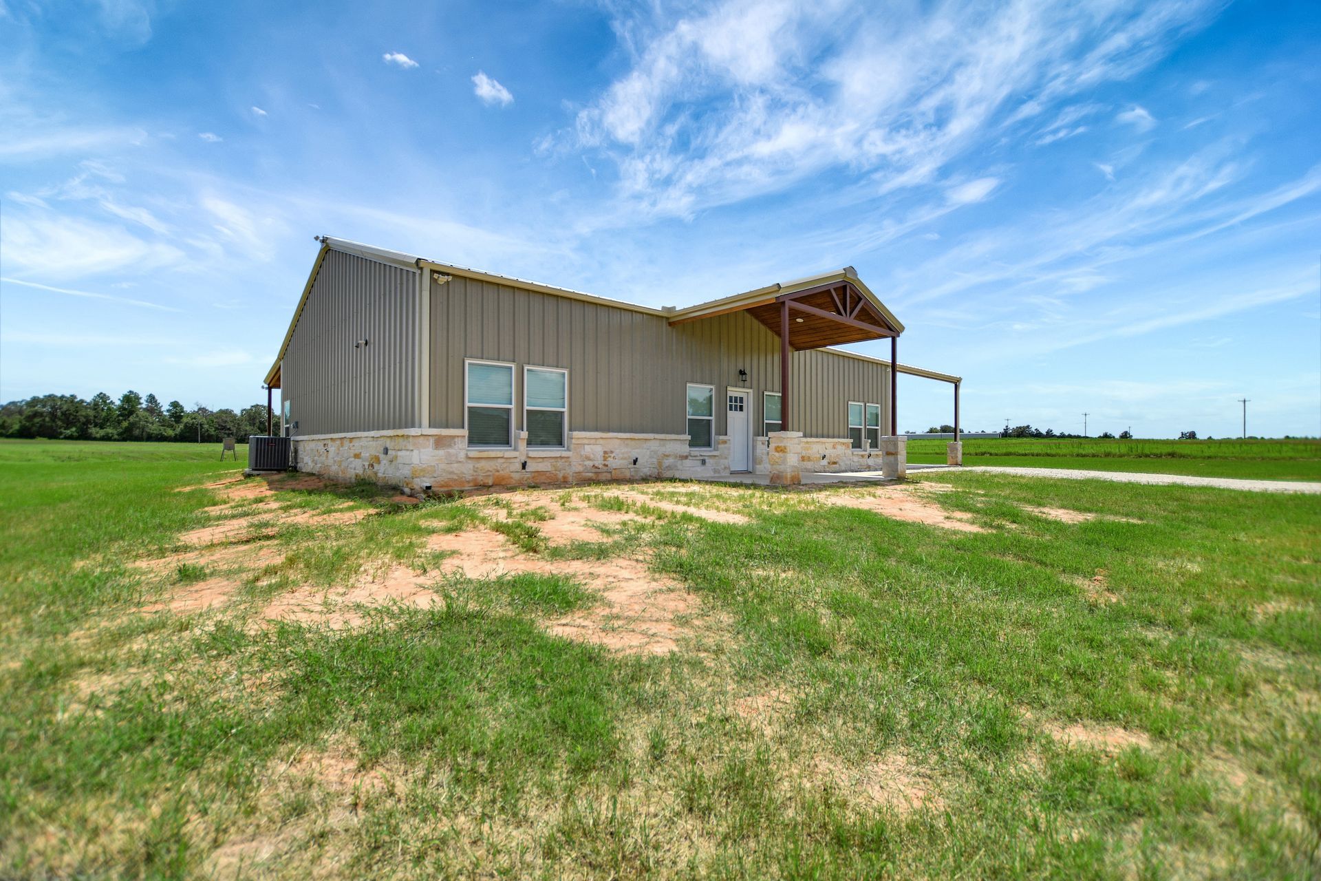 Metal-sided house with covered porch, set on a grassy lot under a blue sky with clouds.