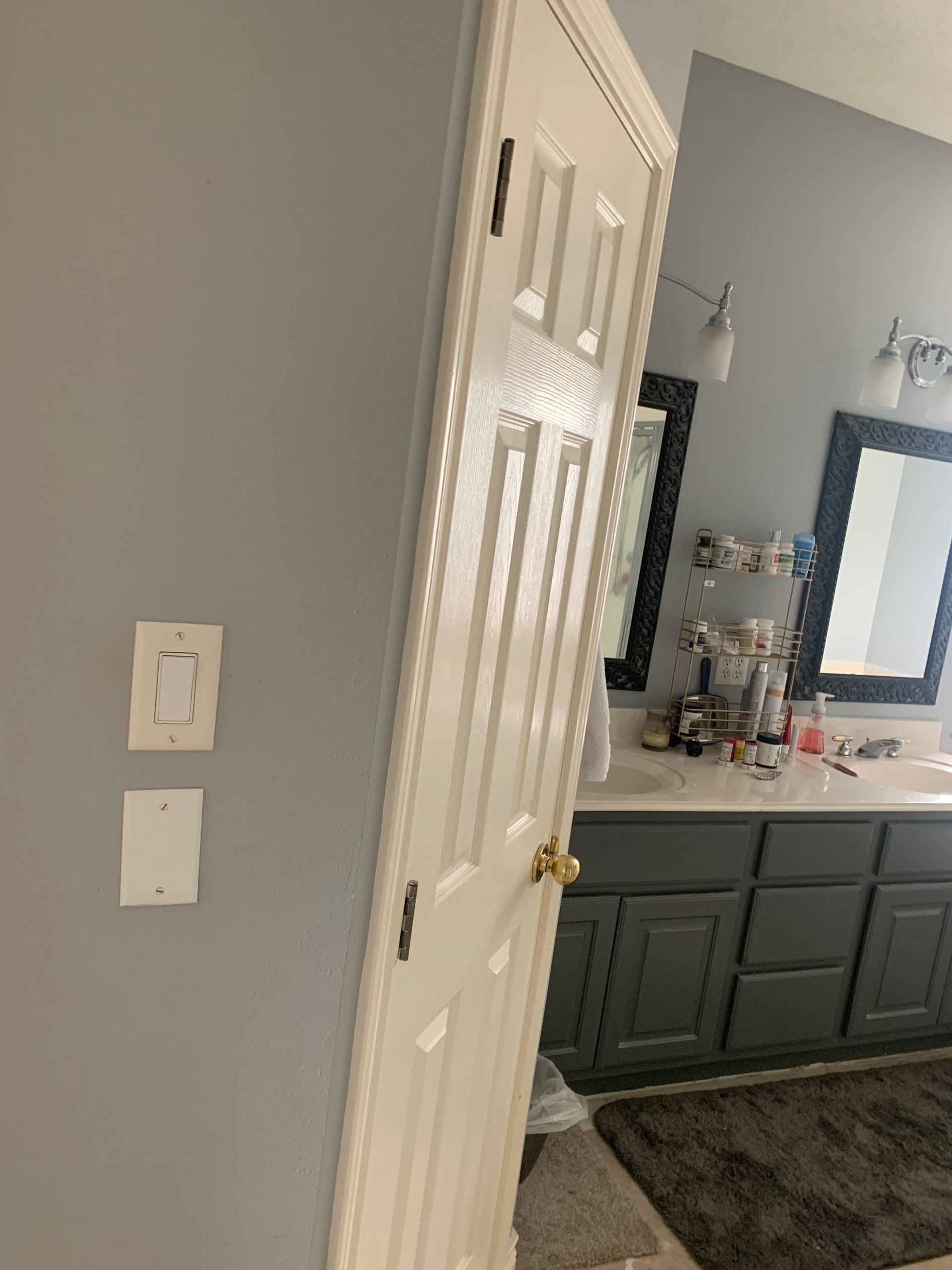 White door slightly ajar in a gray-walled bathroom.  Vanity with two mirrors is visible.
