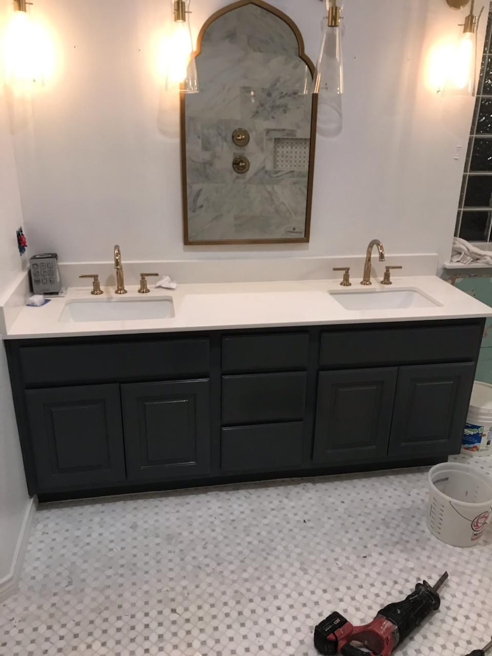 A bathroom with a dark blue vanity, white countertops, gold fixtures, and a decorative mirror above two sinks.