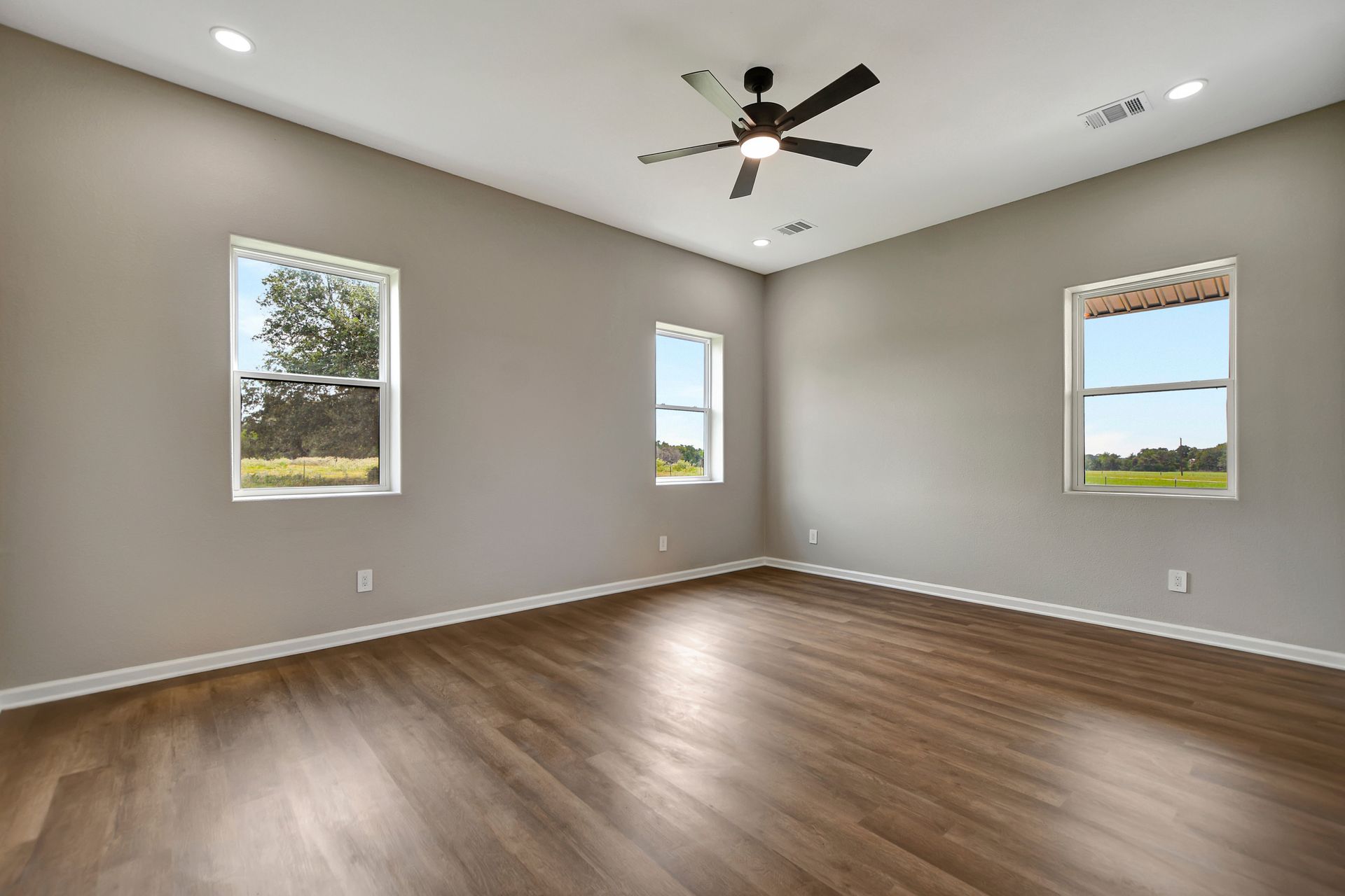 Empty room with wood-look floor, gray walls, and three windows overlooking a green landscape. Black ceiling fan.