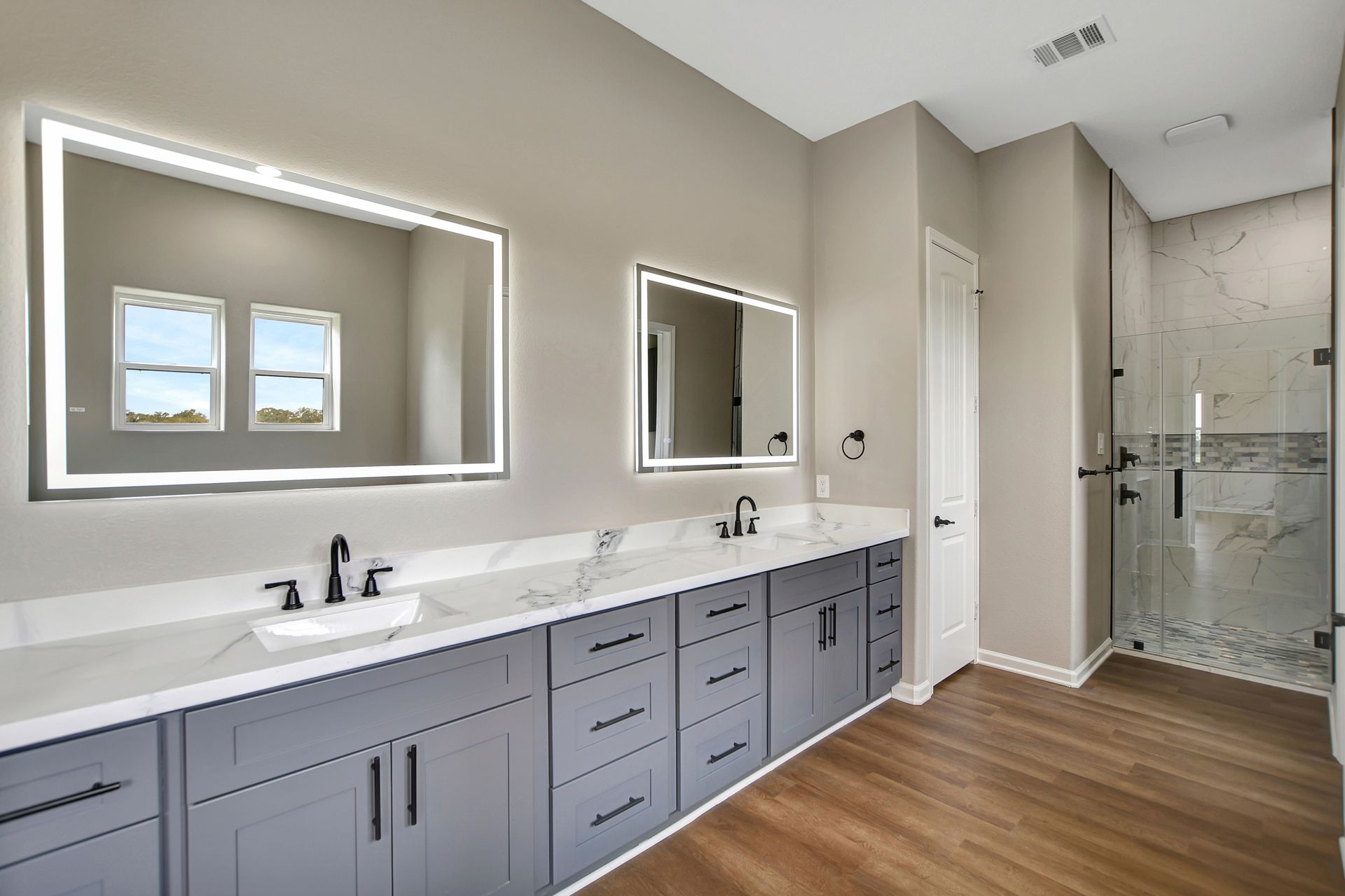 Modern bathroom with gray cabinets, white countertops, two sinks, mirrors, and a glass shower.