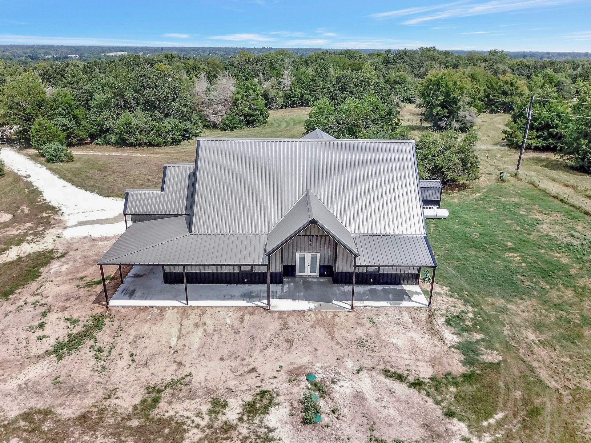 Modern farmhouse with metal roof, porch, and expansive green surroundings under a blue sky.