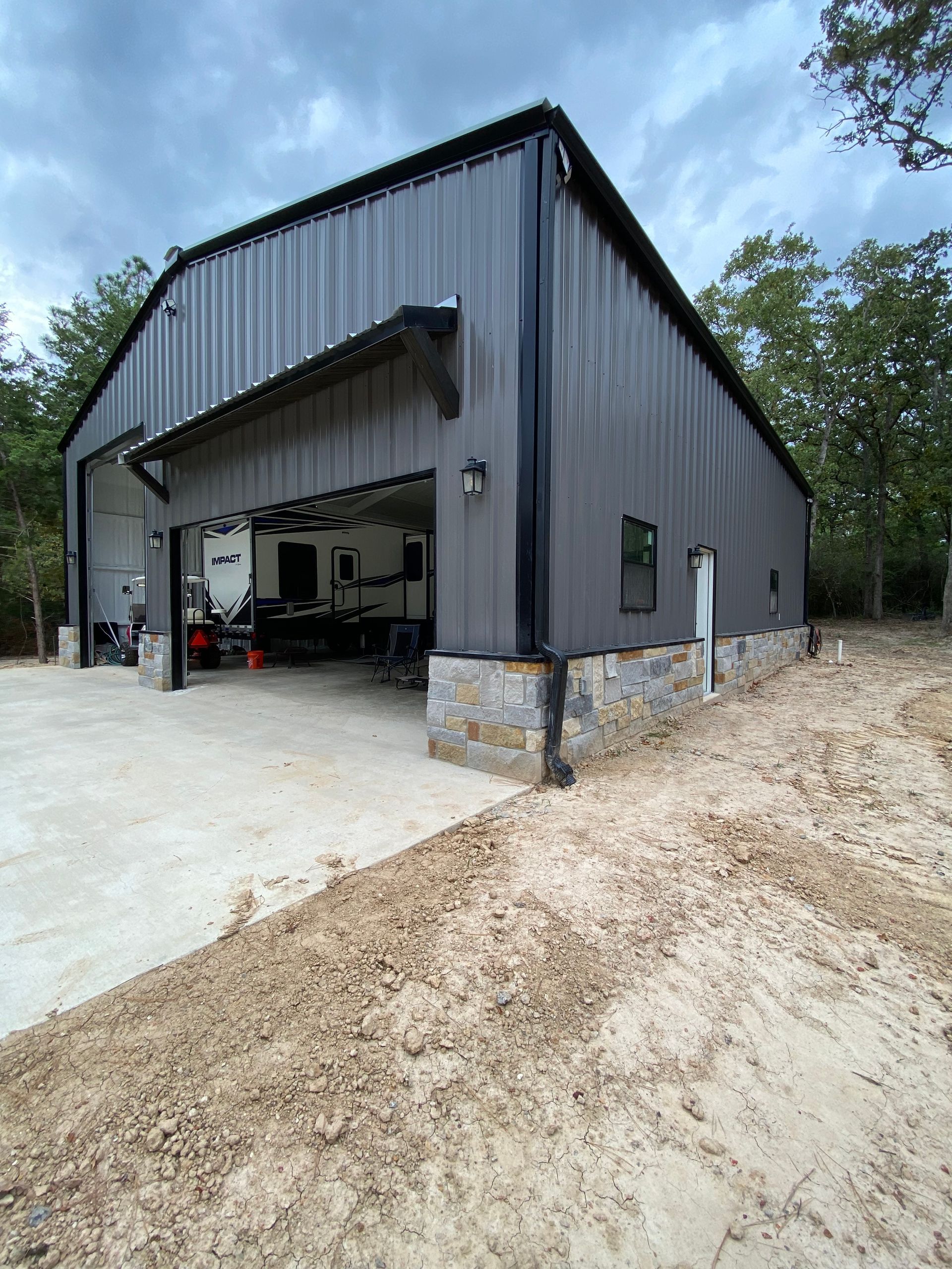Gray metal building with stone base, open garage door, concrete pad, and trees.