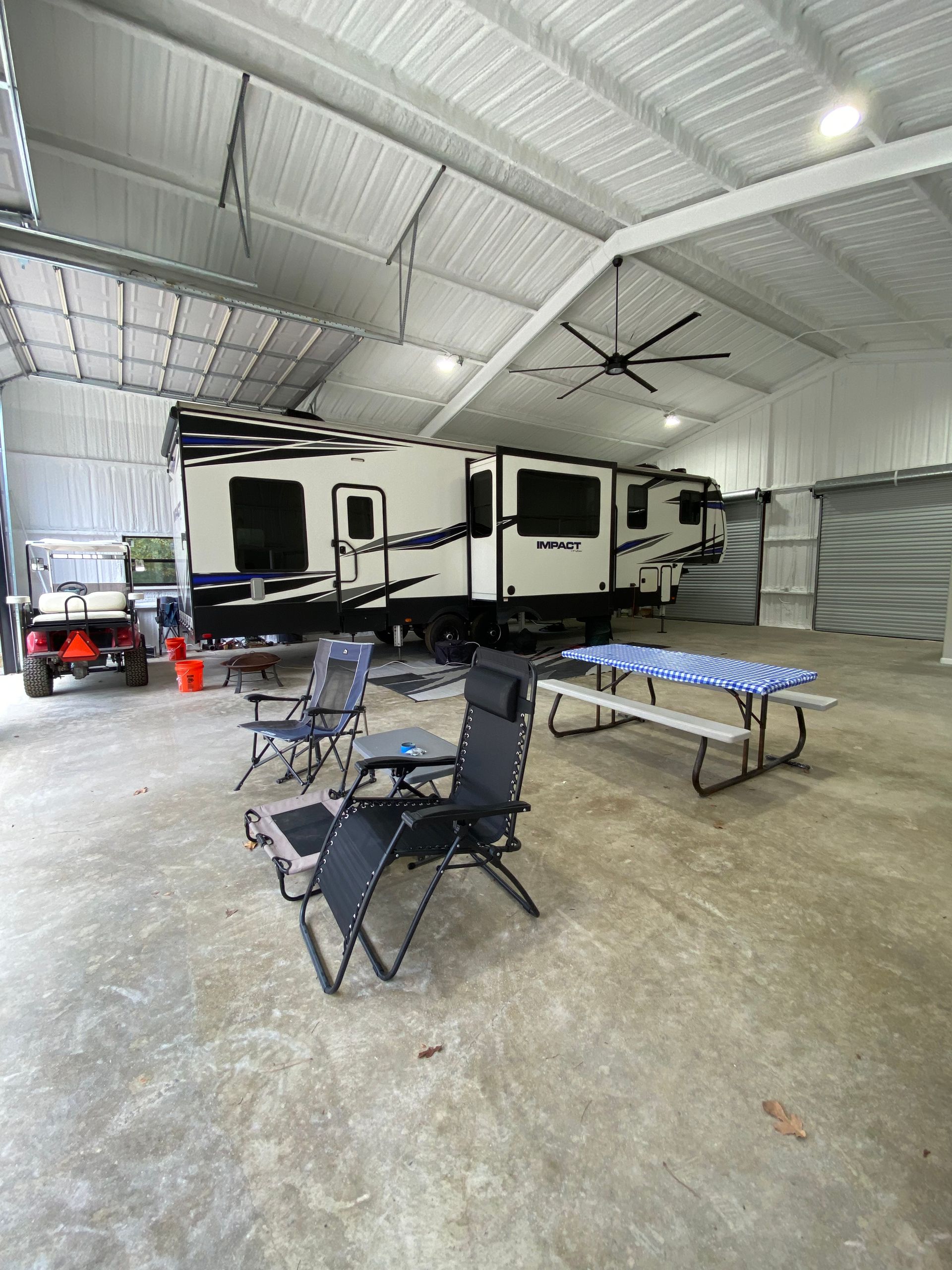 RV parked inside a large metal building. Chairs, a picnic table, and a golf cart are nearby.