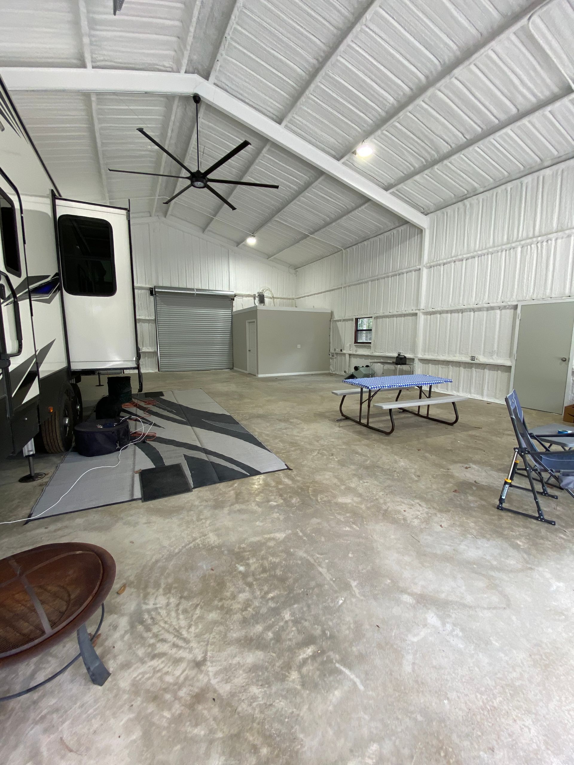 RV parked inside a large white metal building with picnic table and chairs; a fire pit sits in the foreground.