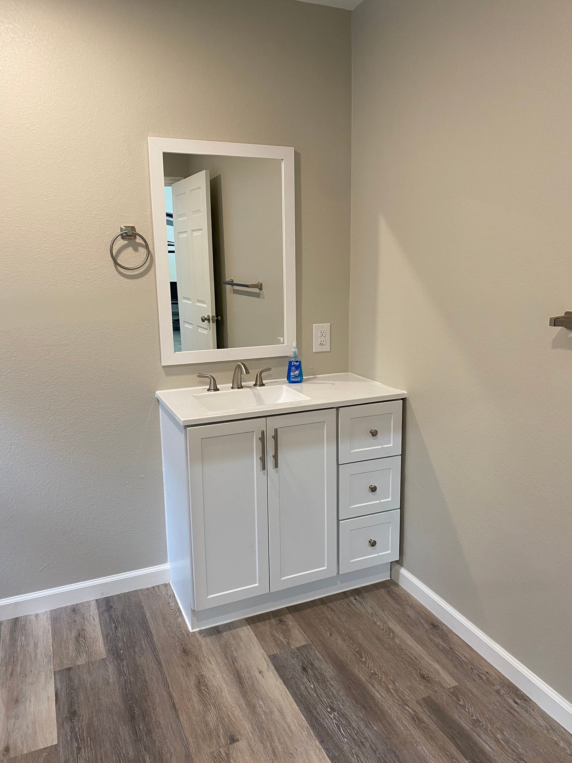 White bathroom vanity with mirror and drawers, mounted on light wood-look flooring, in a room with beige walls.