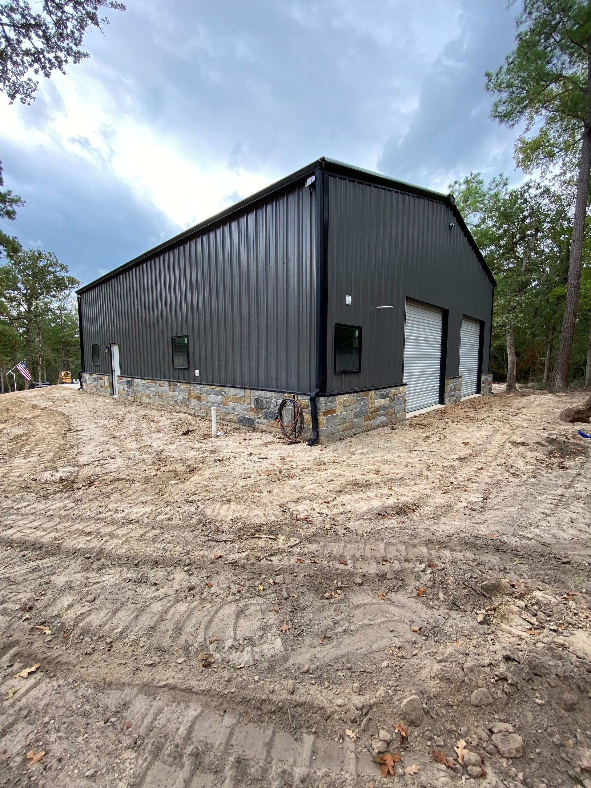 Dark gray metal building with stone base and two garage doors, set in a sandy construction site.