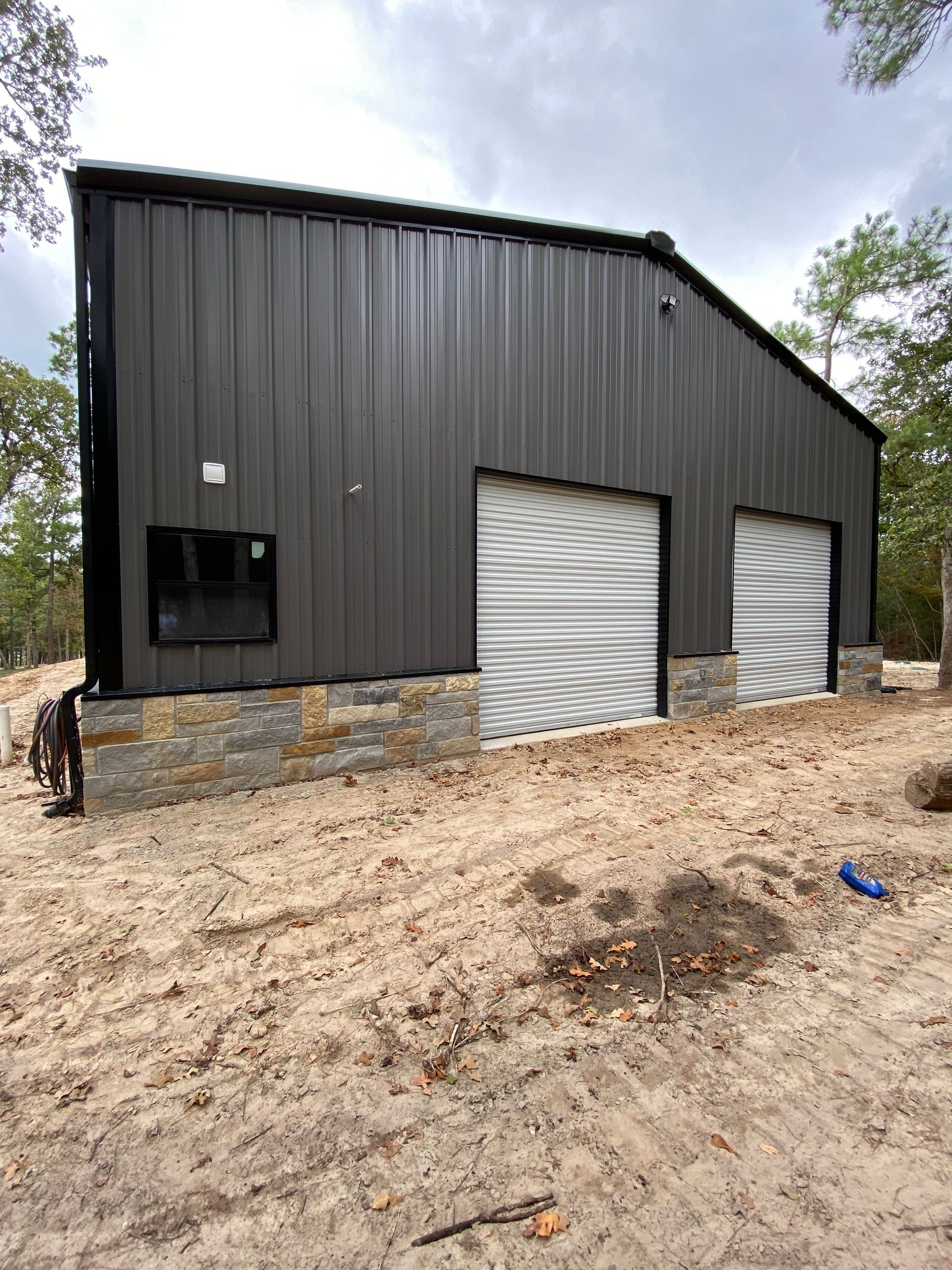 Dark gray metal-clad building with stone base, two garage doors, and small window in a dirt lot.