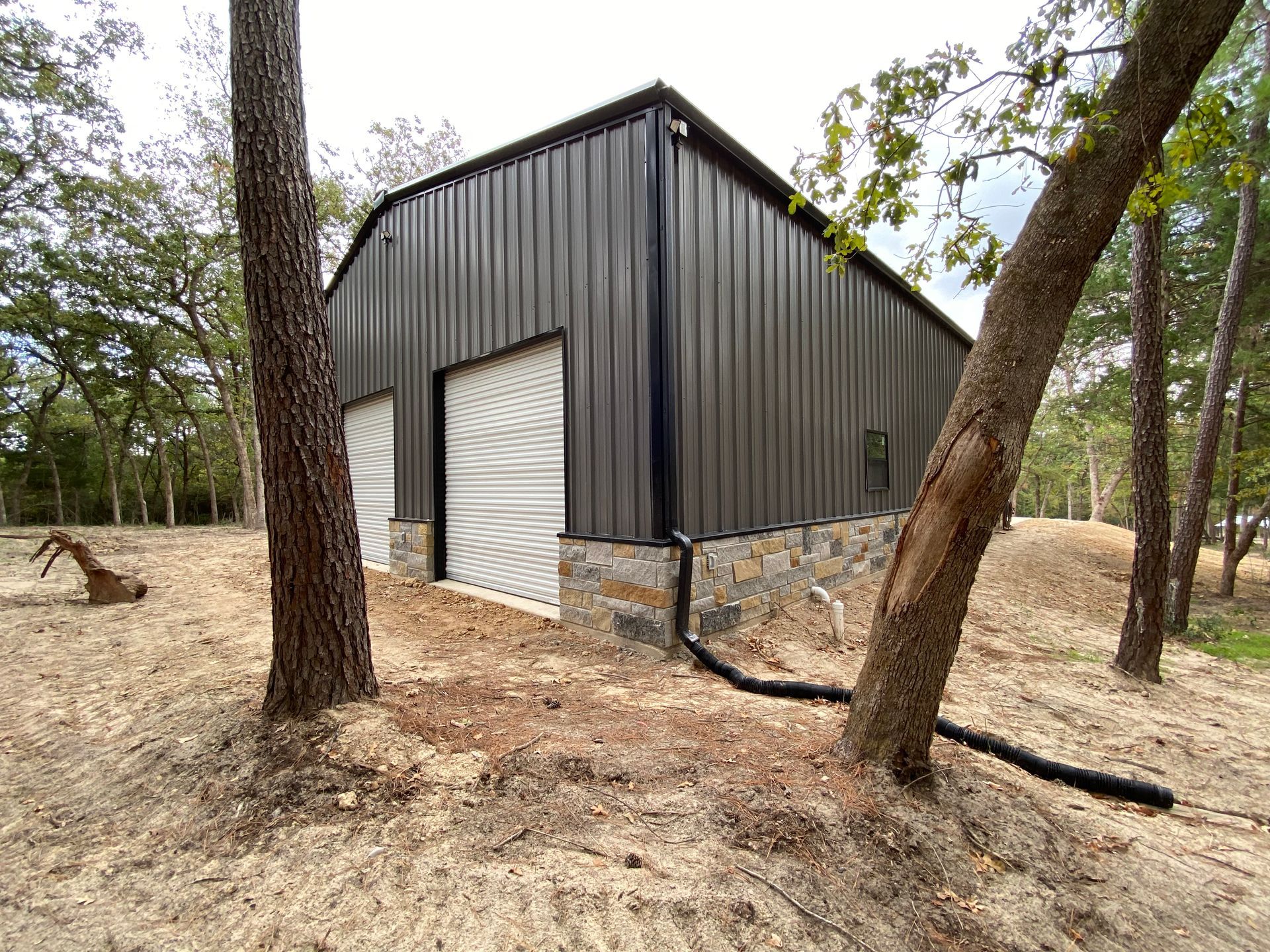 Dark gray metal barn with stone base, set amongst trees on sandy ground.