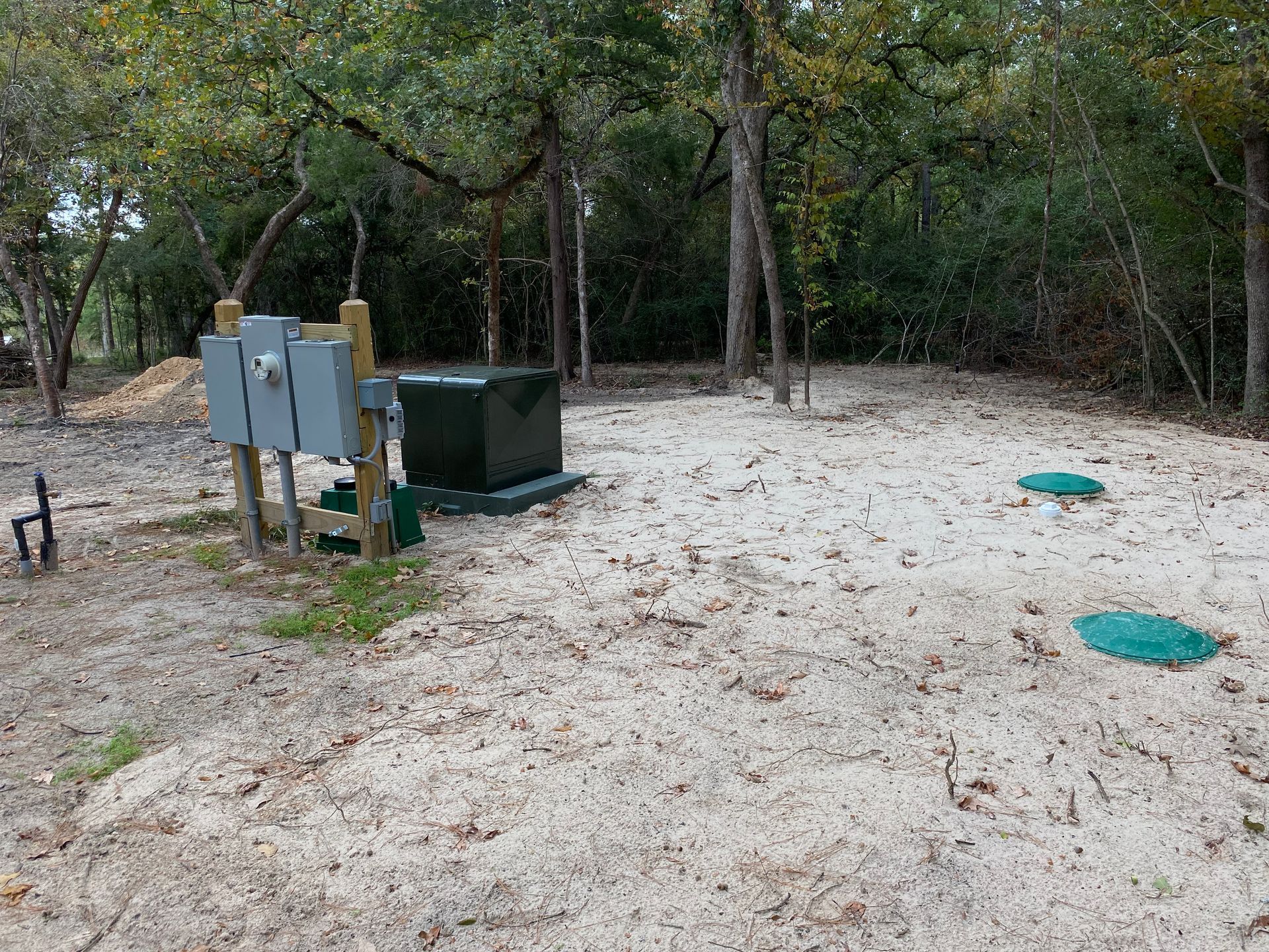 A sandy area with septic system components and electrical boxes, surrounded by trees.