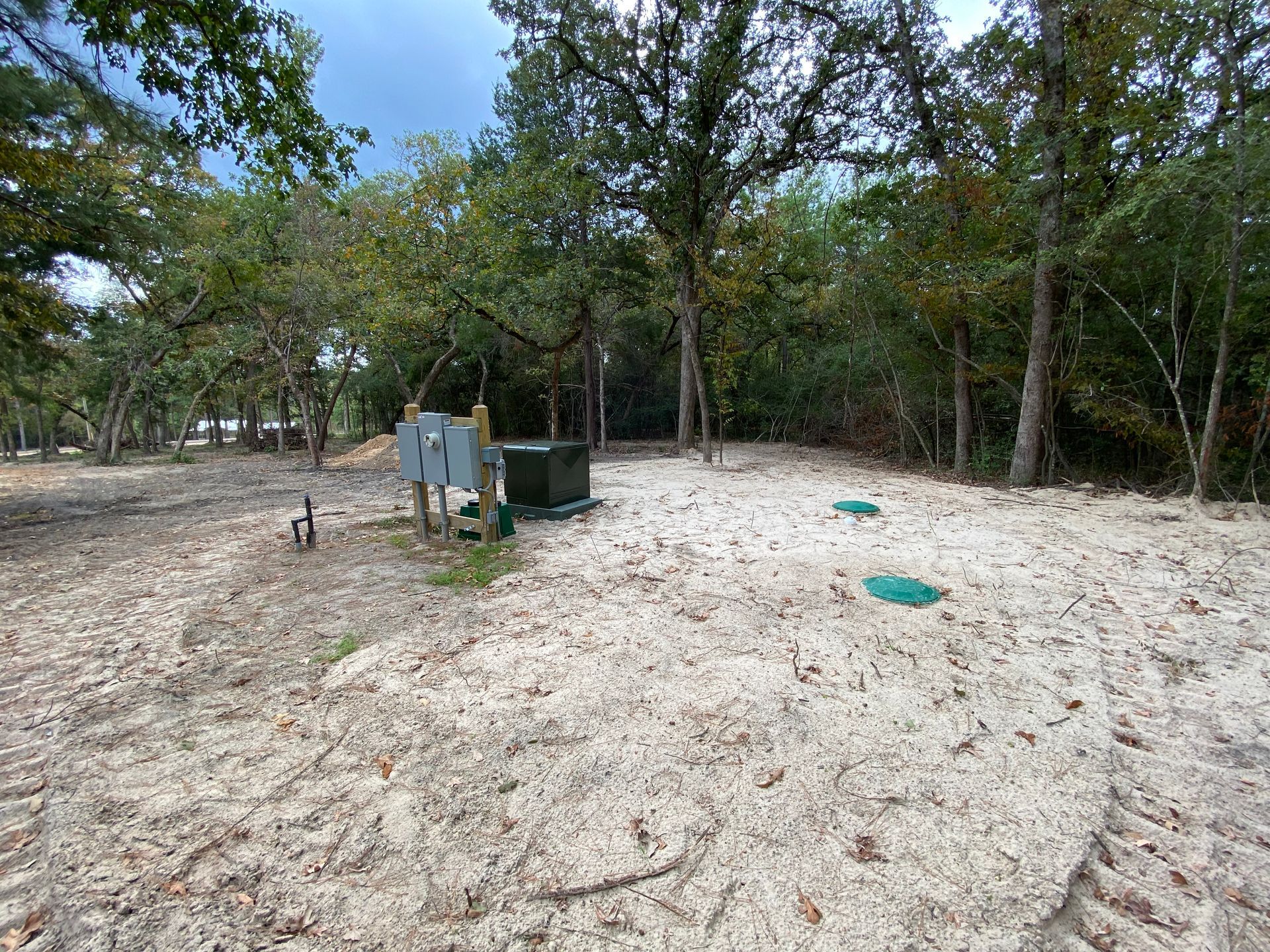 Sandy clearing with trees, a green utility box, and two green circular lids.