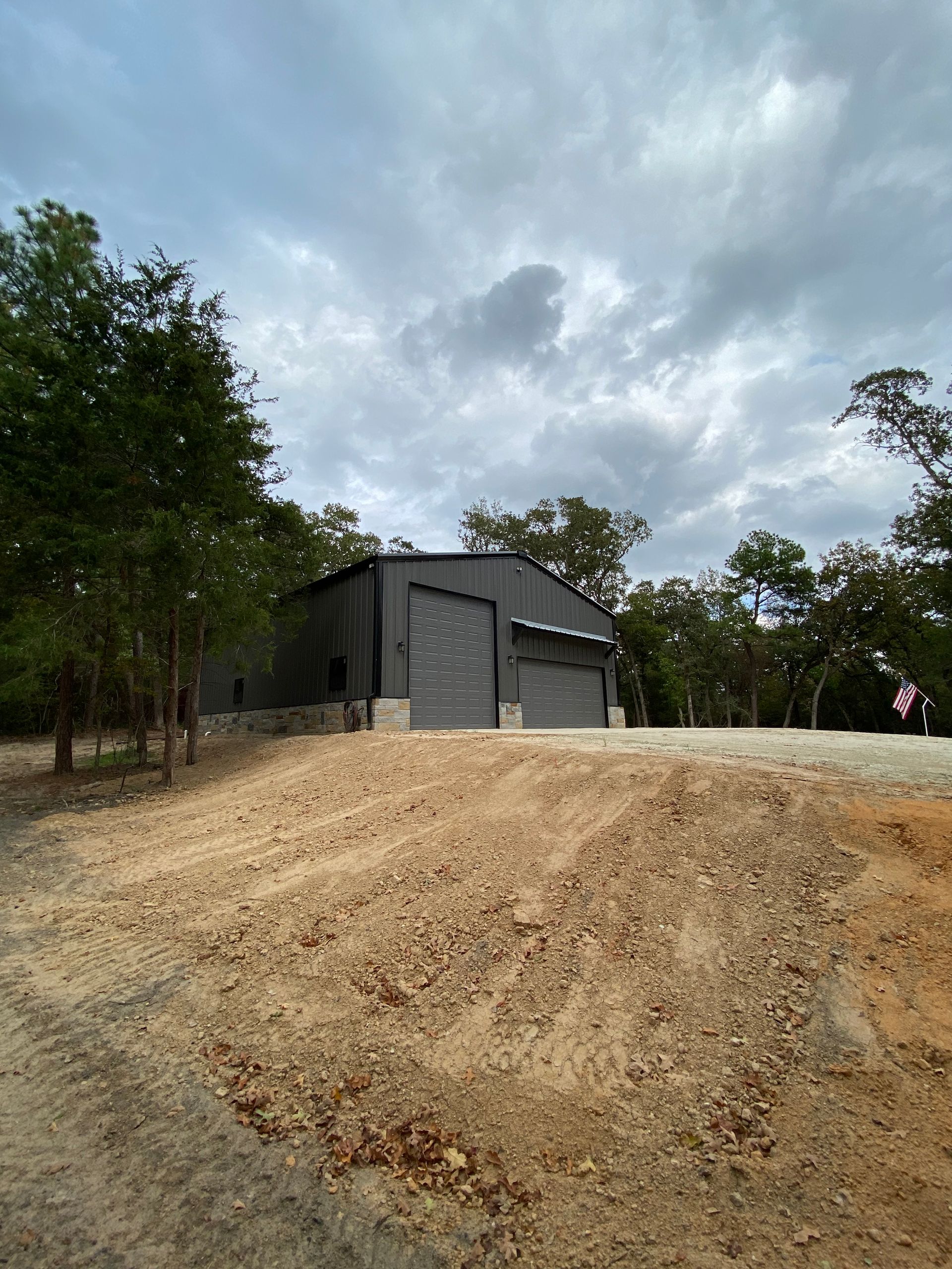 Dark gray metal building with two garage doors on a dirt path under a cloudy sky.