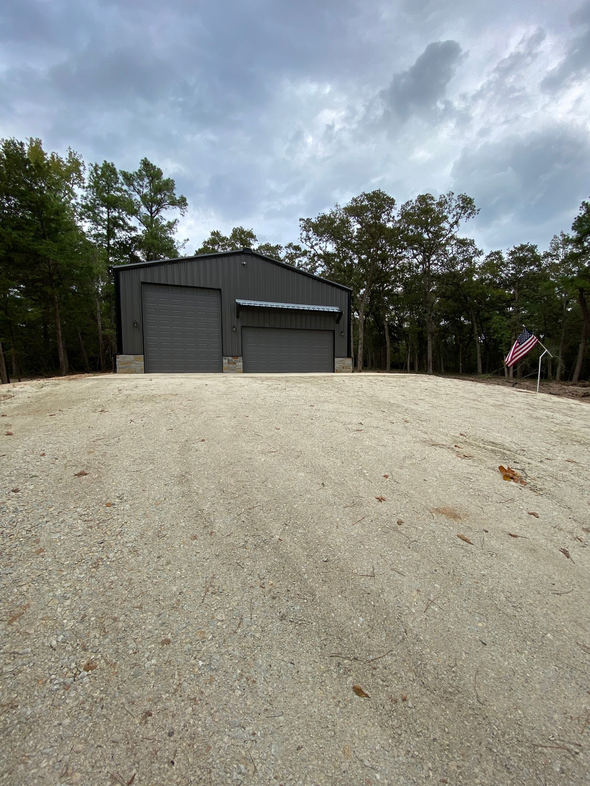 Dark grey metal building with two garage doors on a gravel drive, cloudy sky.
