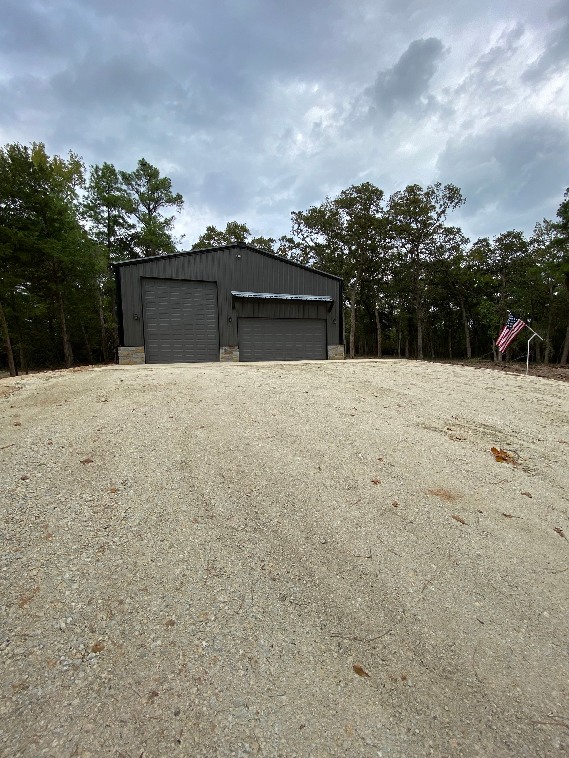 Gray metal building with two garage doors on gravel driveway, surrounded by trees.