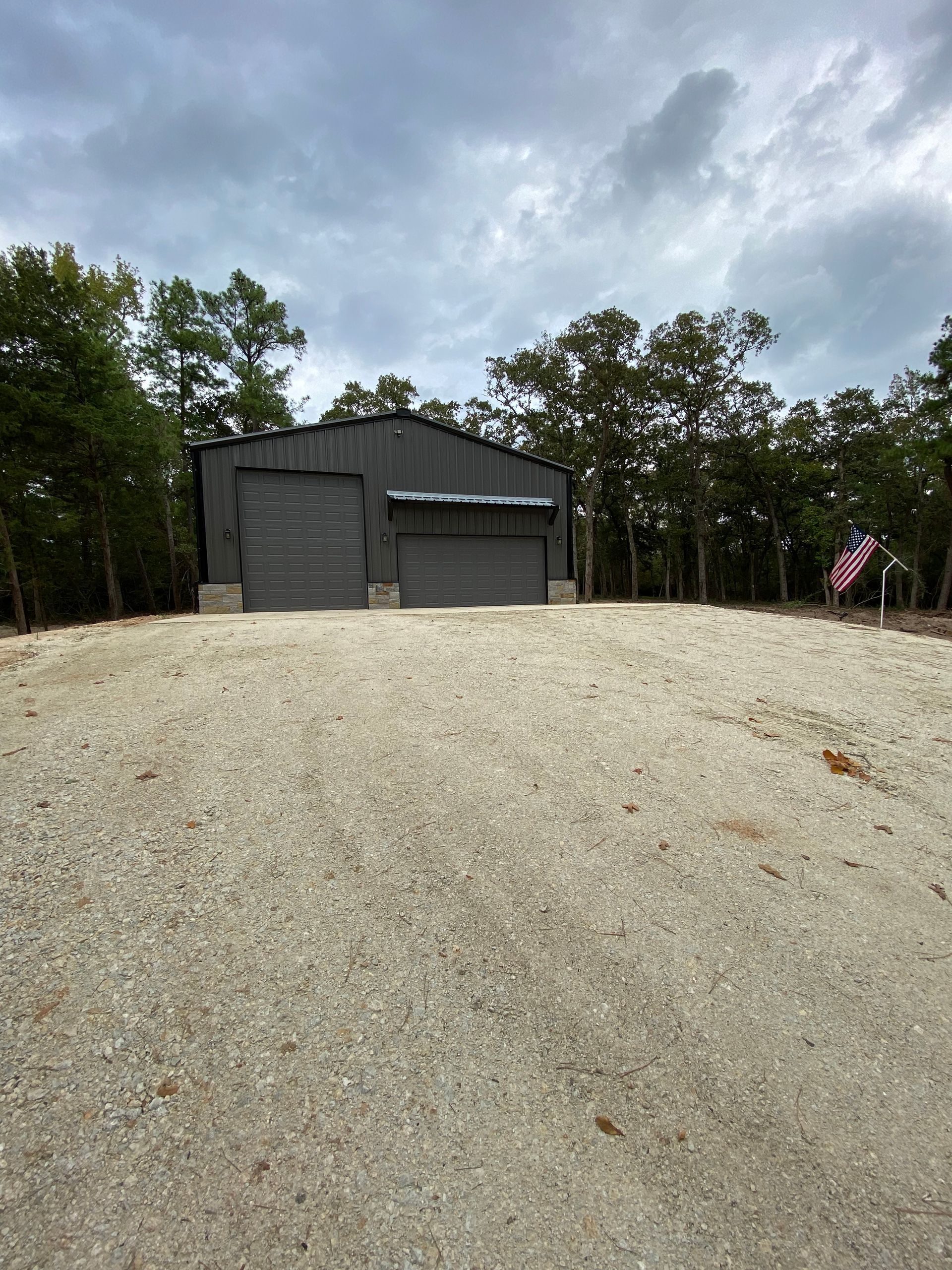 Gray metal garage with gravel driveway, cloudy sky, surrounded by trees.