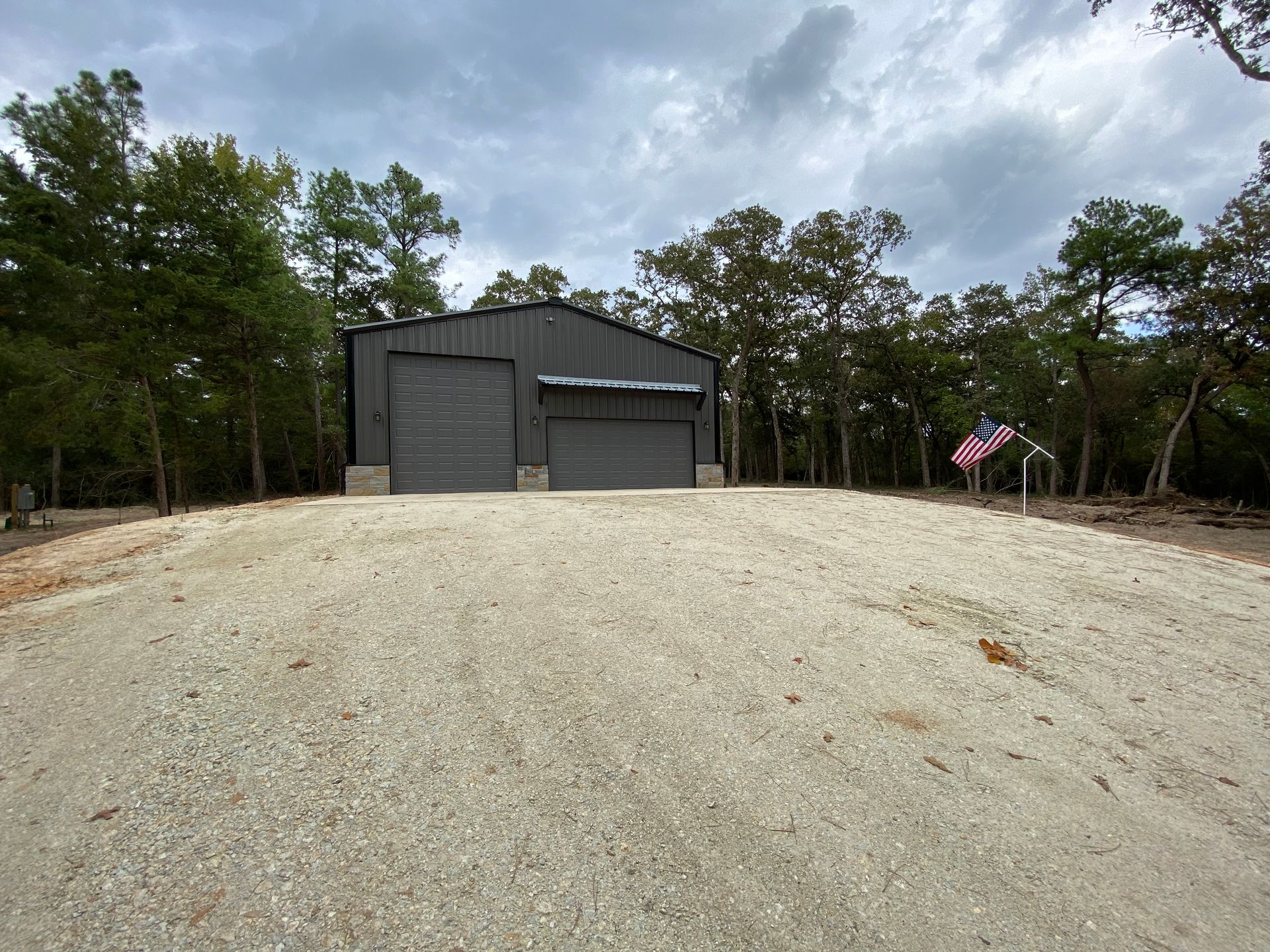 Gray metal building with two garage doors on a gravel lot, American flag.