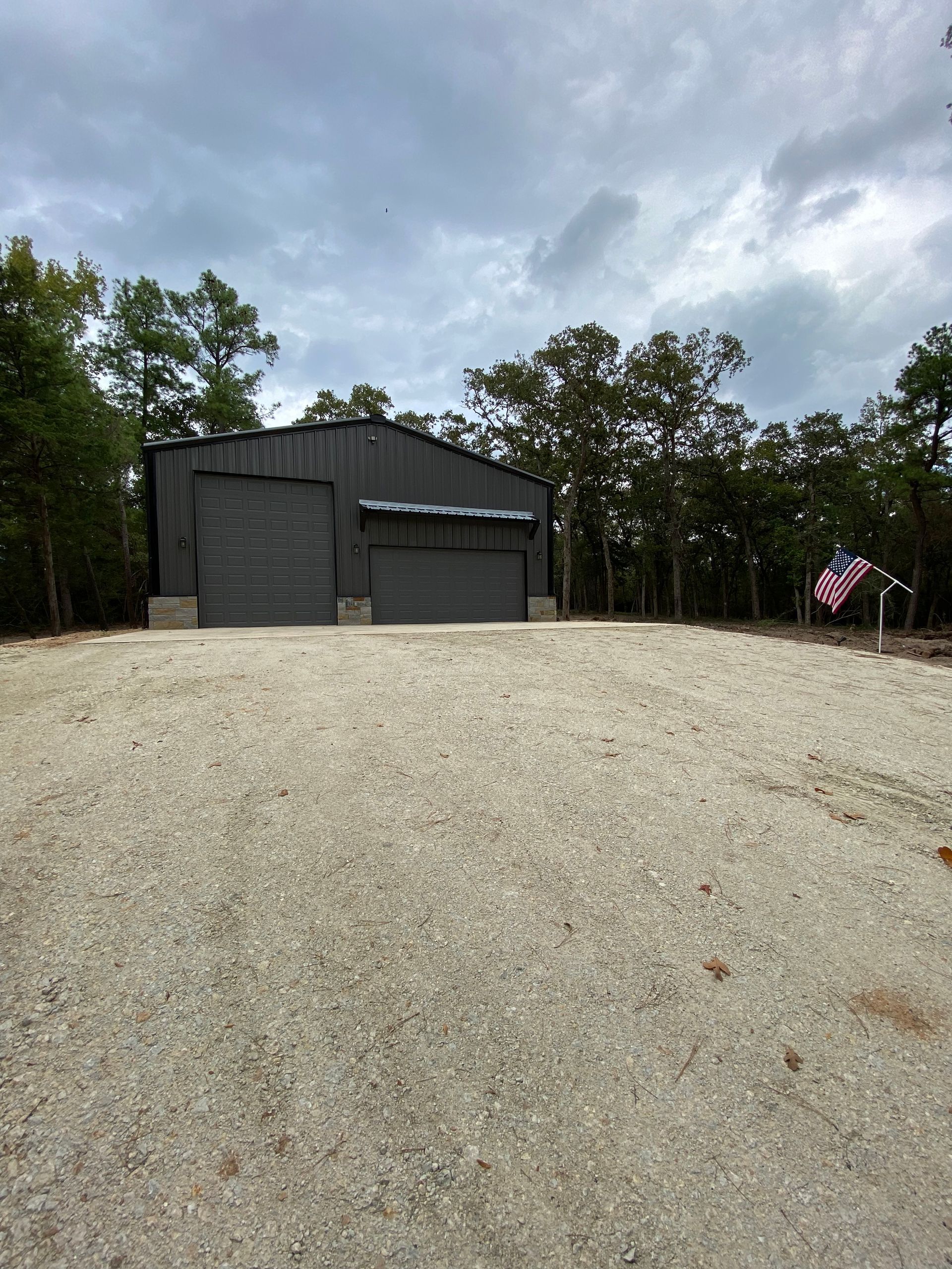 Gray metal storage building with two garage doors, gravel driveway, trees, and cloudy sky.