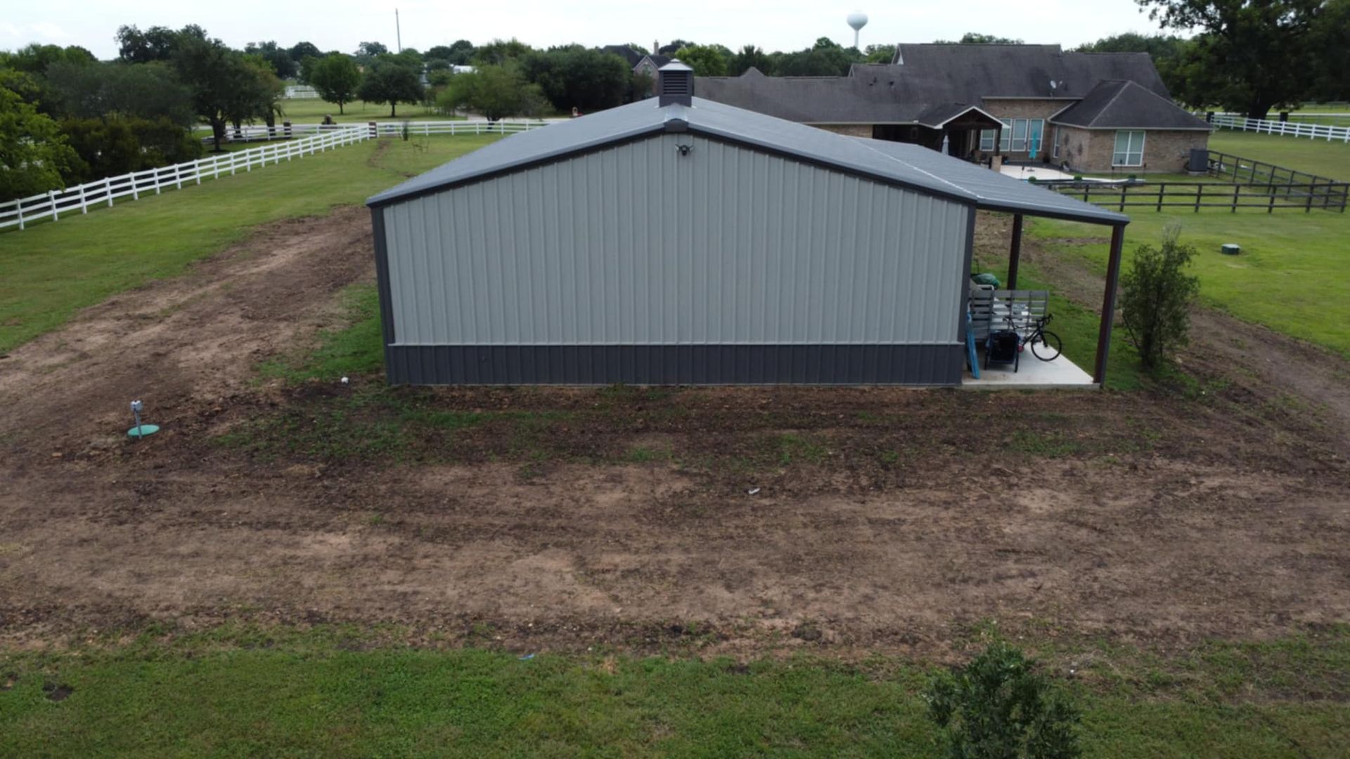 A metal barn with a dark gray bottom and a tan upper portion sits on a grassy hill next to a house.