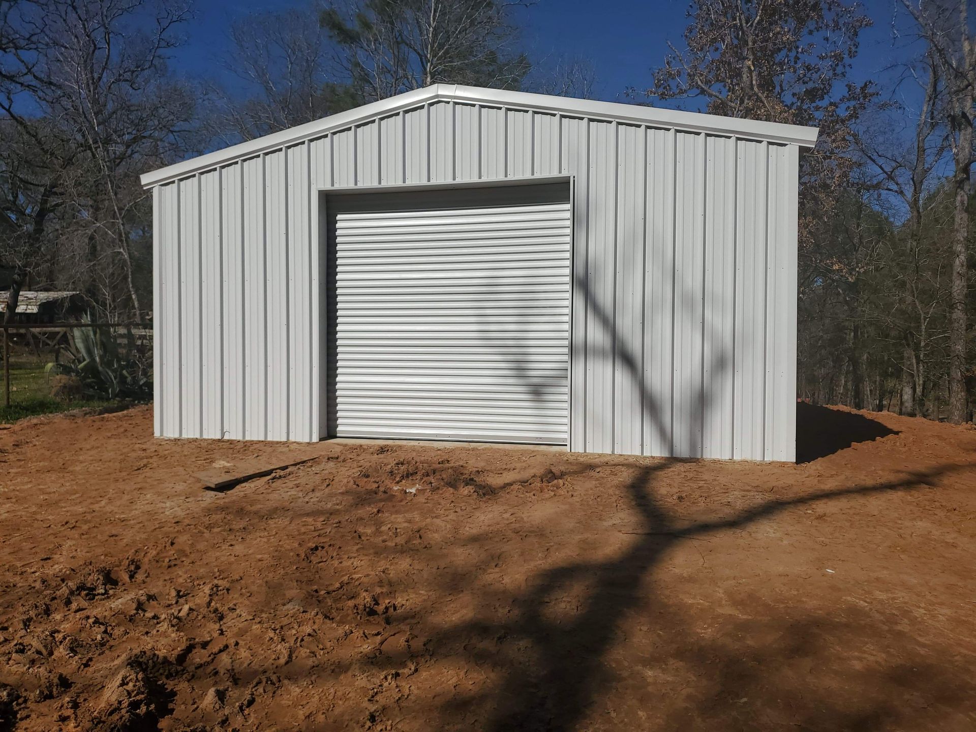 White metal shed with a roll-up door, standing on a dirt lot, surrounded by trees.