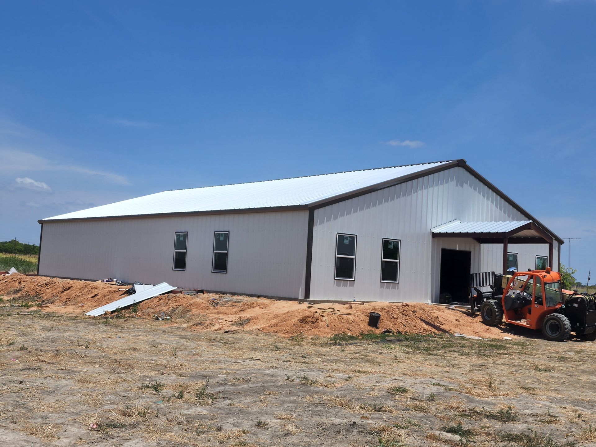 White metal building under construction on a sunny day, surrounded by dirt and a small construction vehicle.